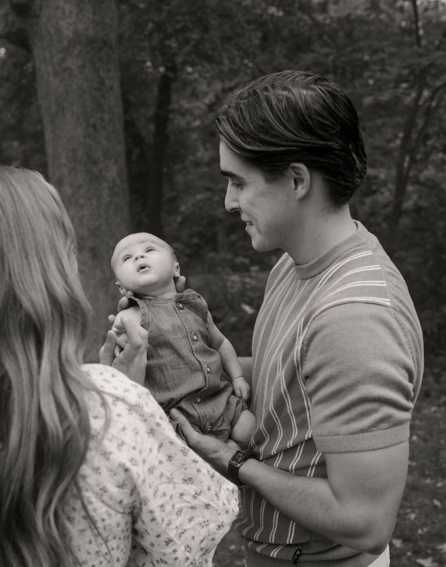 Black and white photo of parents holding newborn baby outdoors among trees.