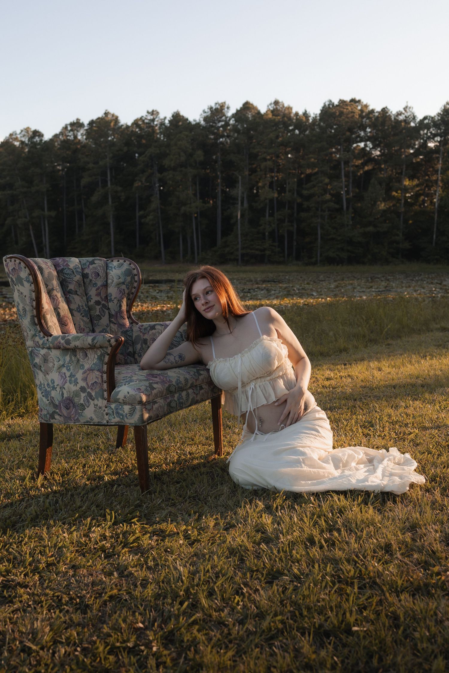 Young woman in a white dress sitting beside a vintage floral armchair in a grassy field at sunset.