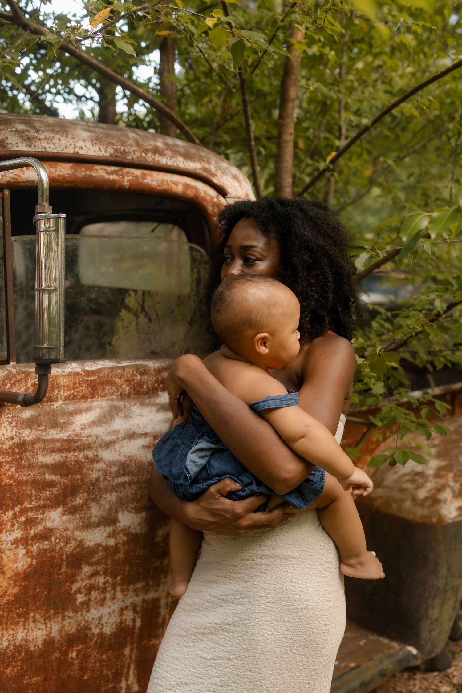 A Black mother in a white dress tenderly embraces her baby beside a weathered rusty vintage truck.
