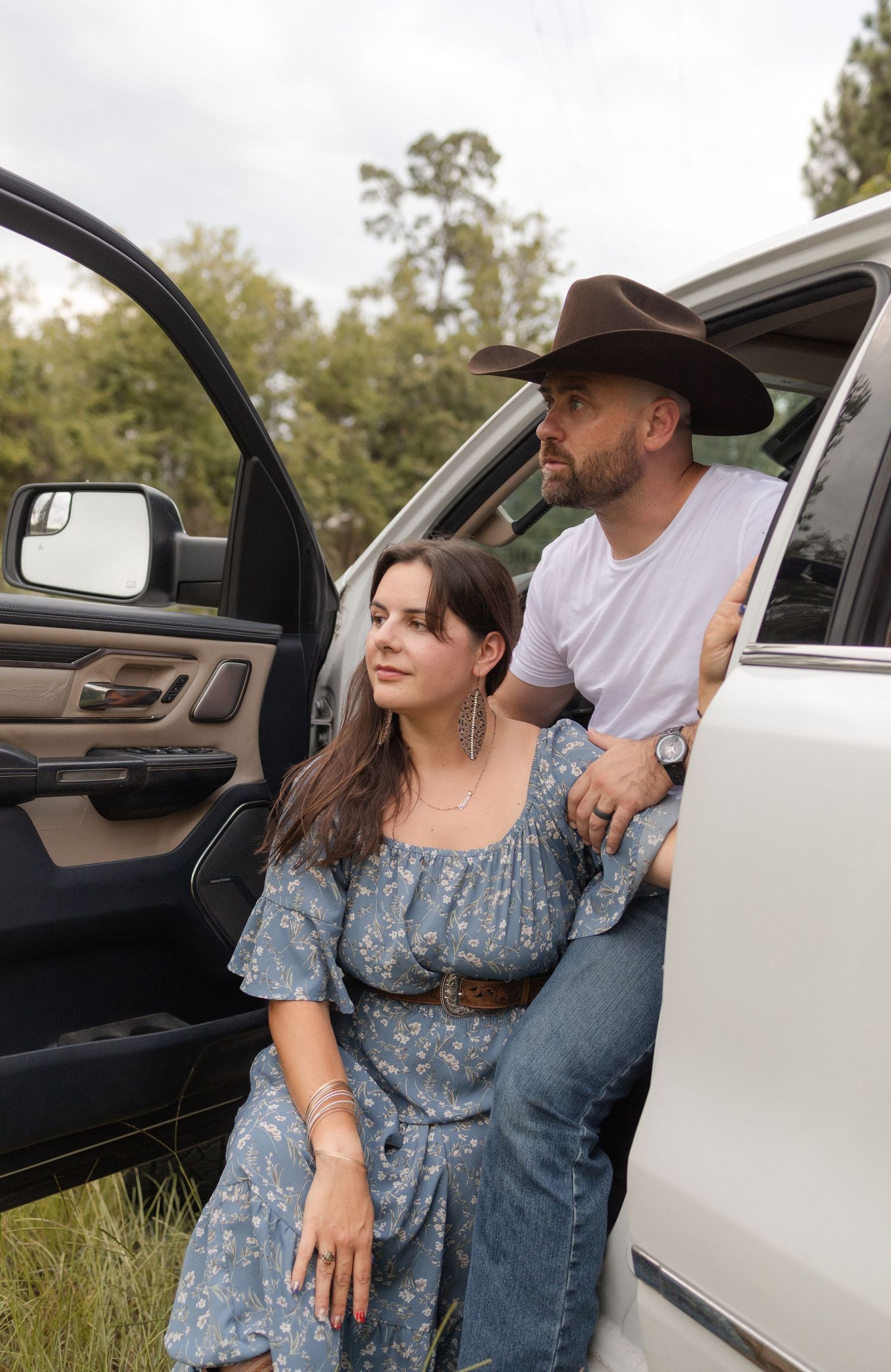 Couple in western wear posing by a white truck in a scenic rural landscape.