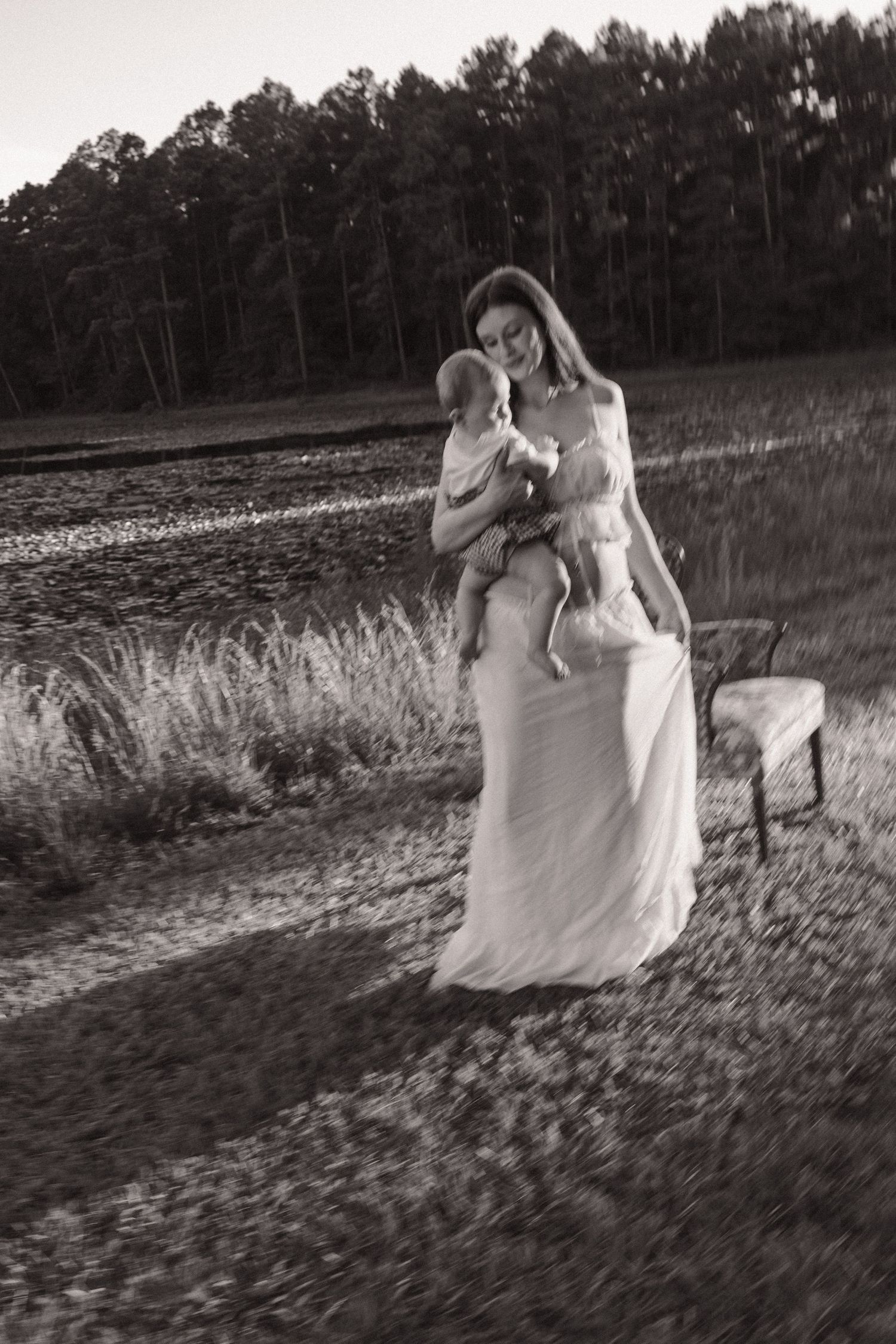Black and white photo of a mother in a flowing white dress holding her baby in an open field at dusk.