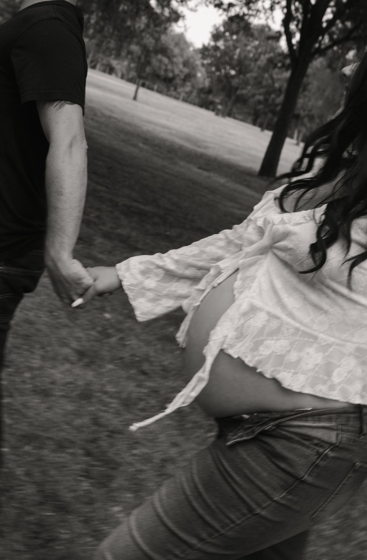 Black and white photo of couple holding hands walking through park, pregnant belly visible.