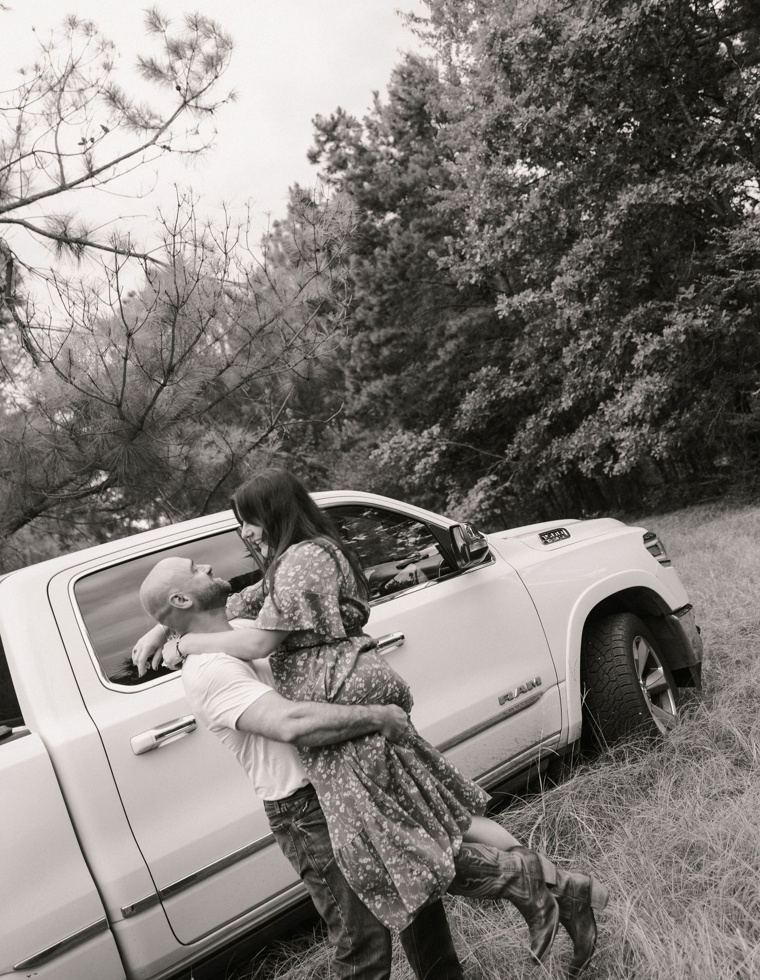 Black and white photo of a man lifting a woman outside a white pickup truck in a wooded area in Fayetteville NC.