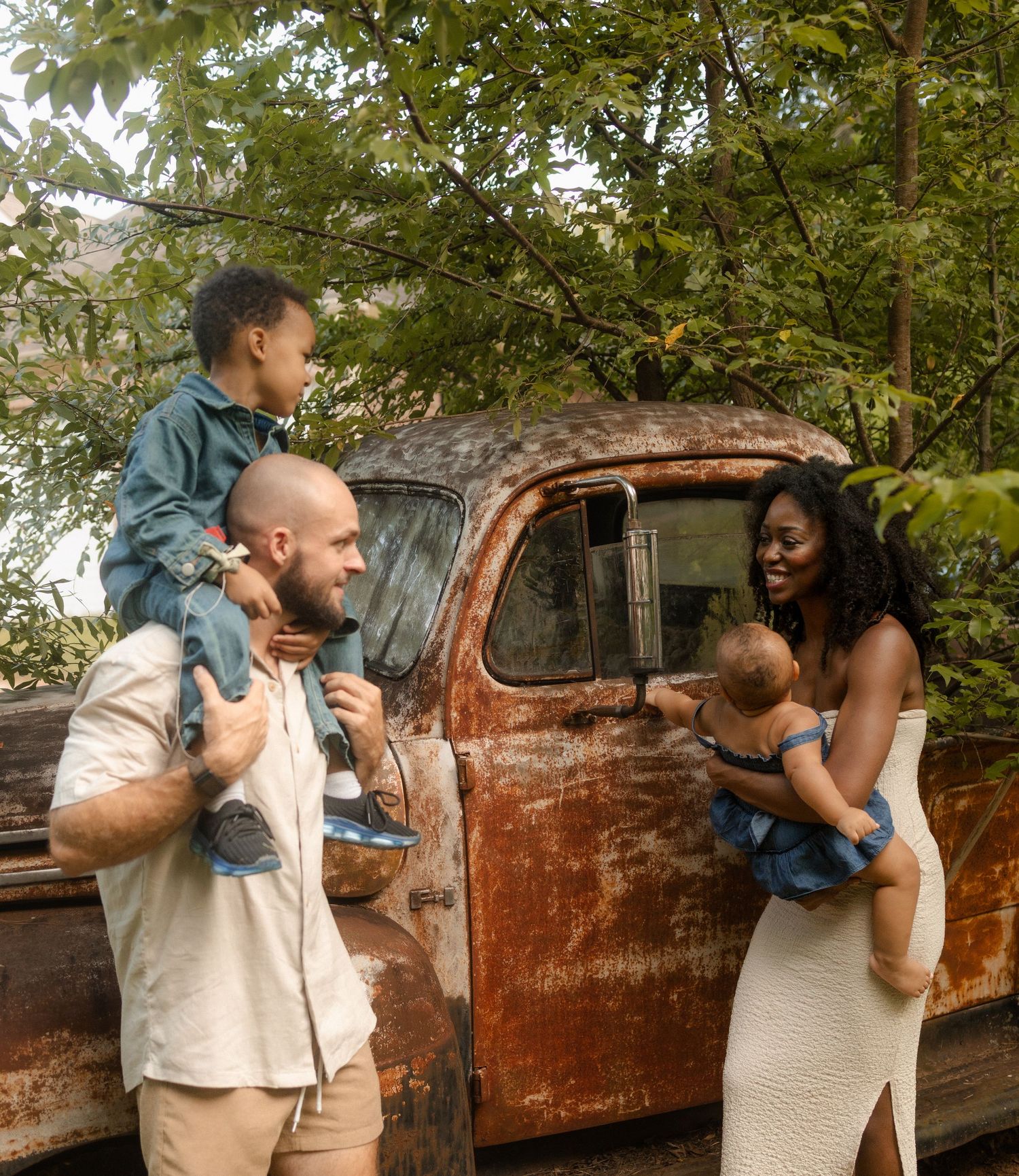 A joyful interracial family poses by a rustic vintage truck outdoors, parents holding two young children.