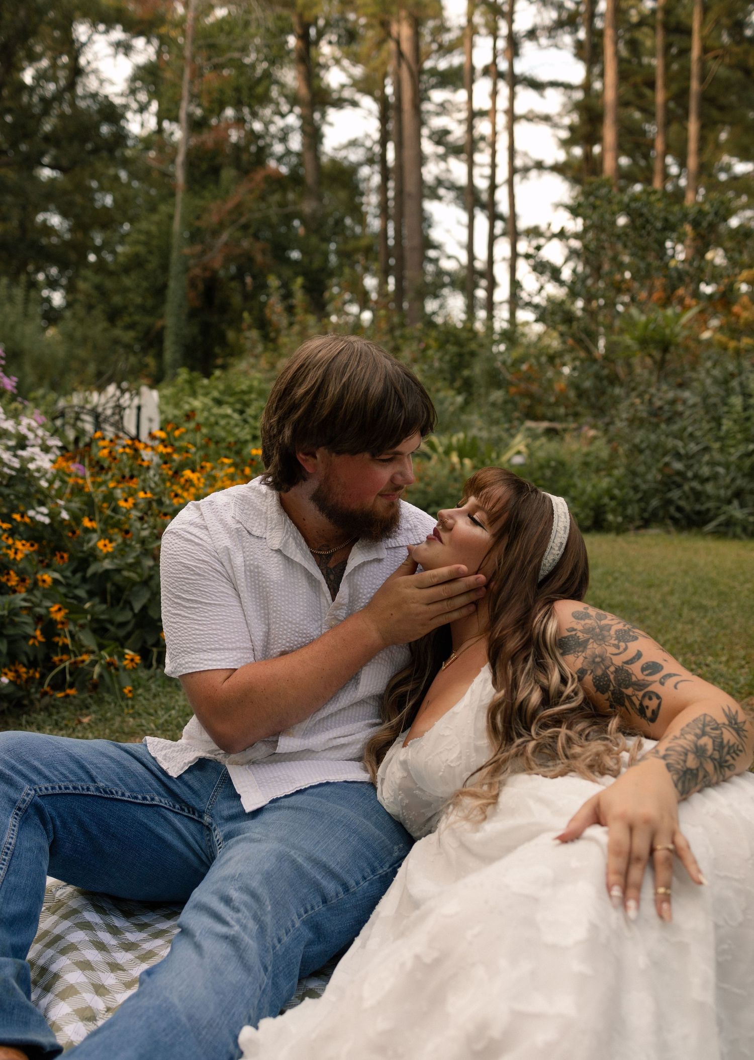 Couple sharing a romantic moment in a garden, groom in jeans holding brides face in a wedding dress.