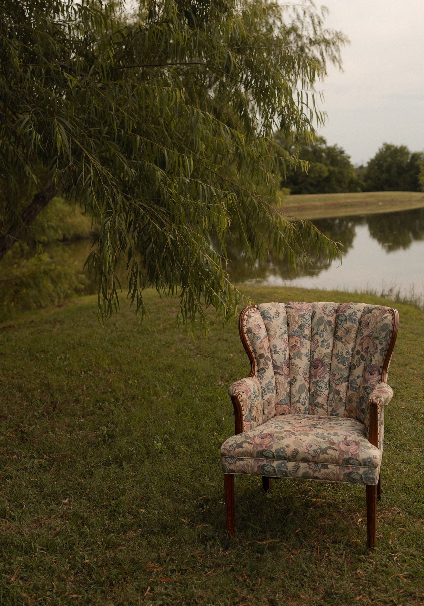 Vintage floral armchair sitting on green grass beneath a weeping willow tree beside a calm reflective pond.