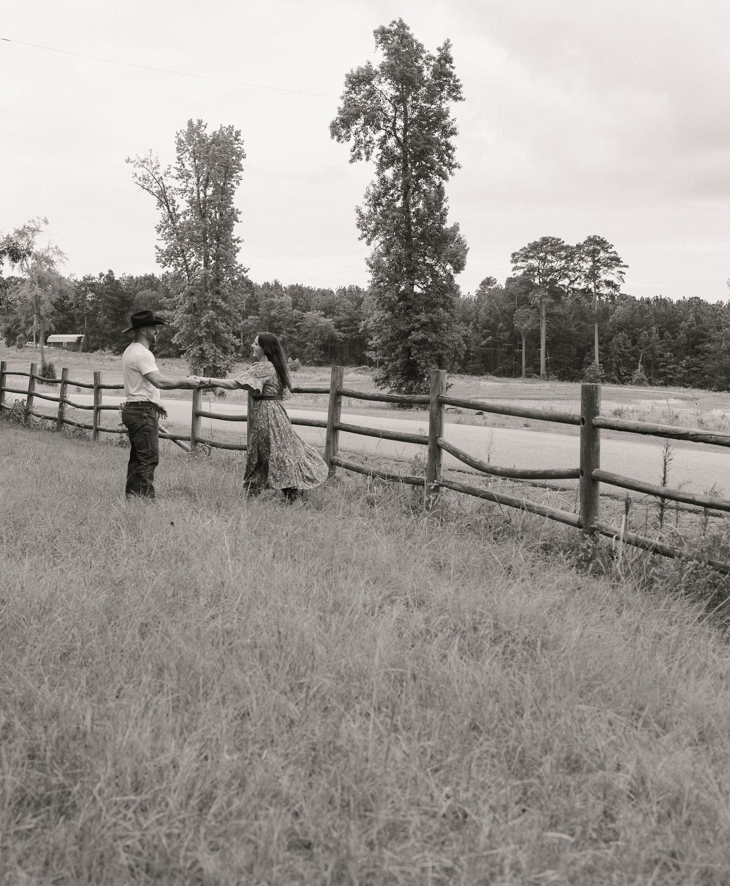 Black and white photo of a couple standing by a wooden fence in a scenic rural field.