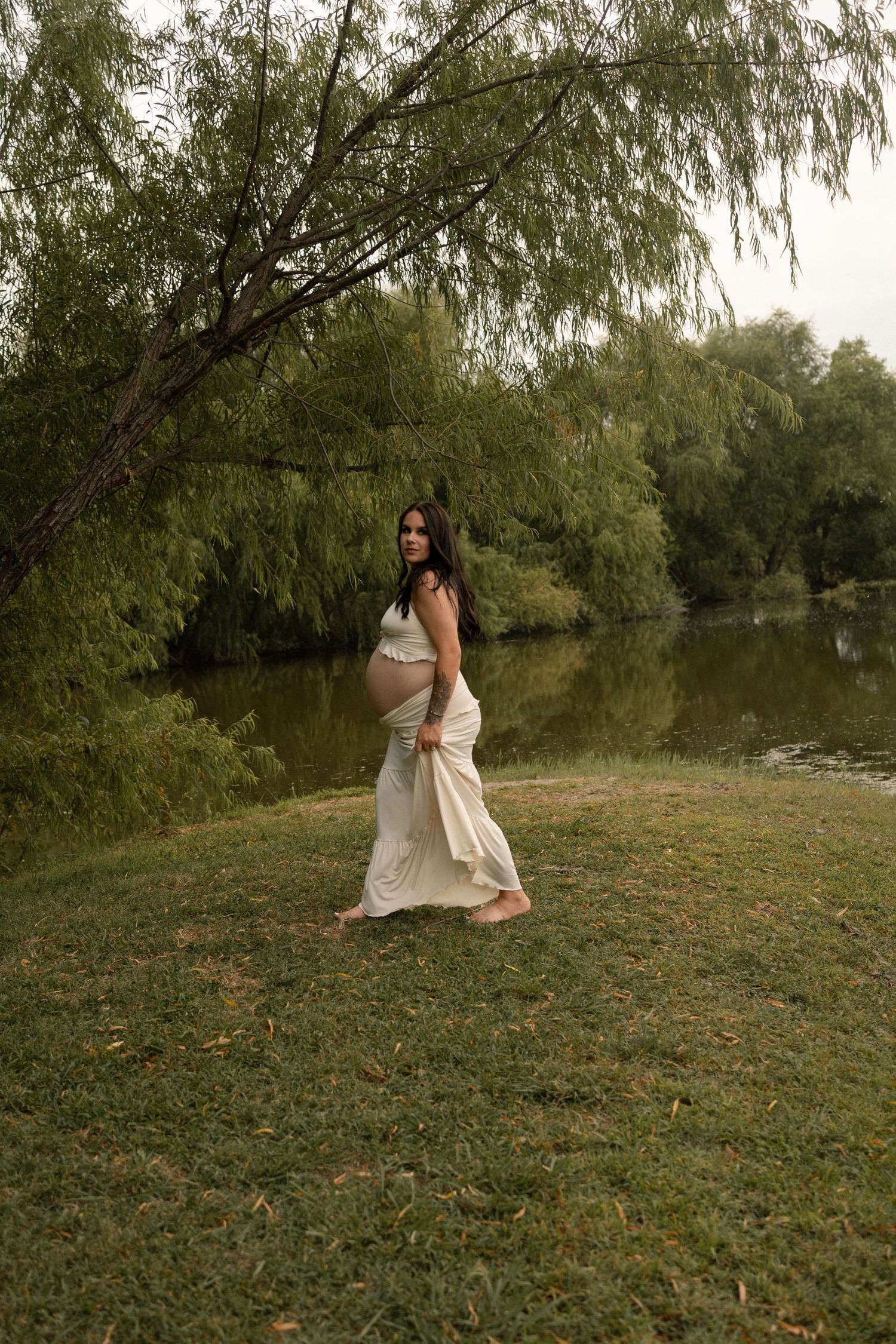 Pregnant woman in white dress walking barefoot by a serene pond under willow trees.
