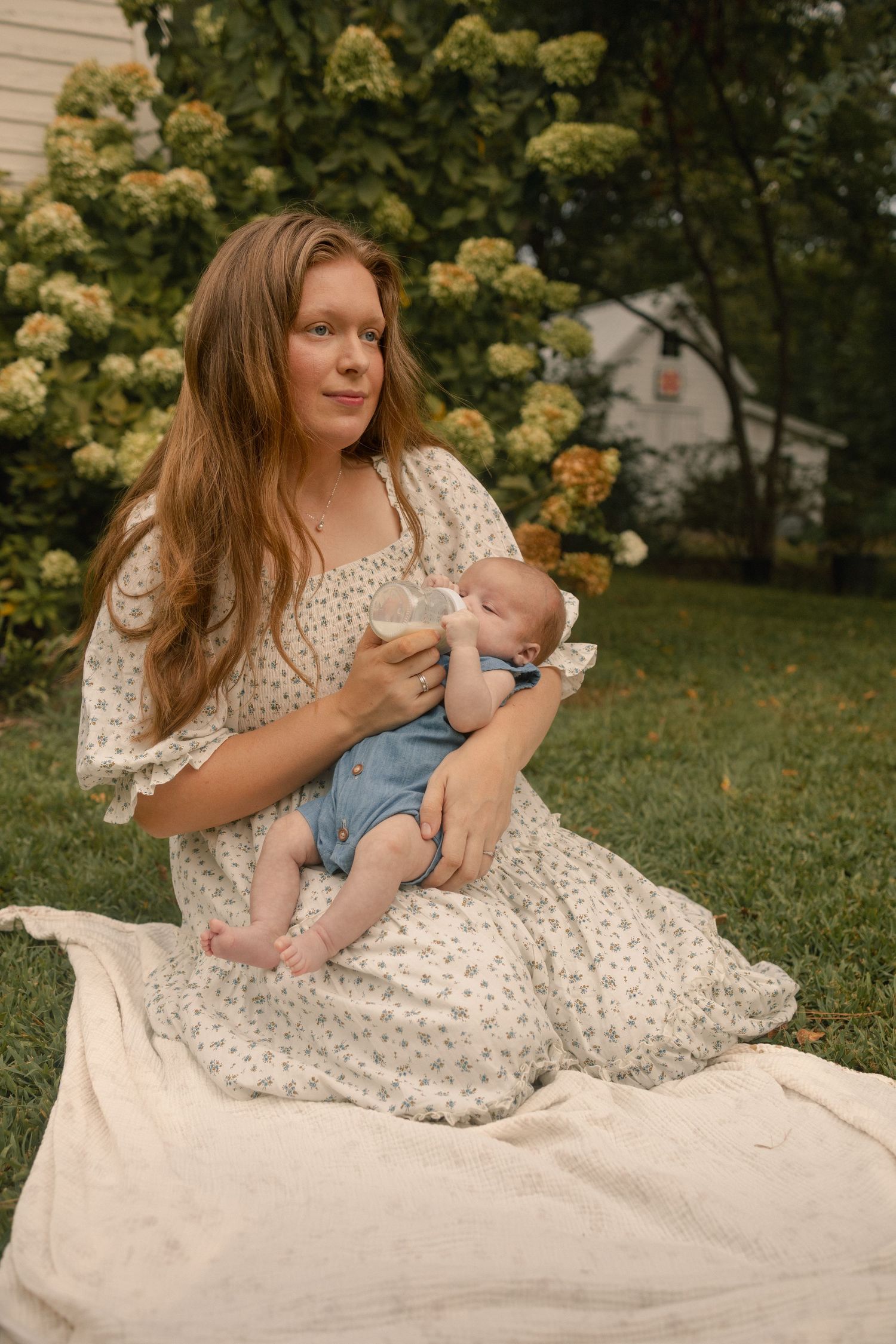 Woman in floral dress bottle feeding newborn baby while sitting on blanket in garden.
