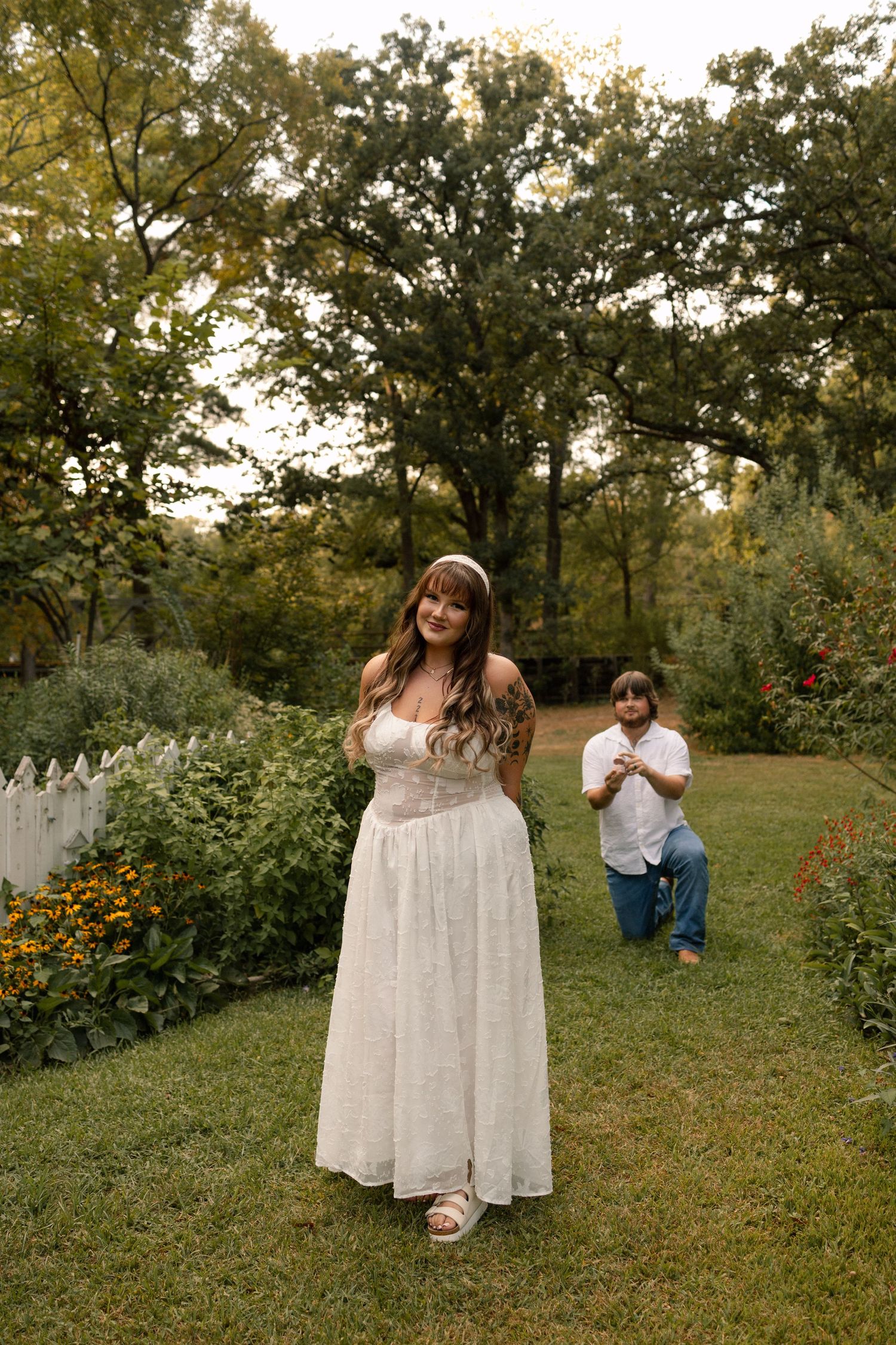 A woman in a white dress smiles as her partner proposes on one knee in a lush garden.