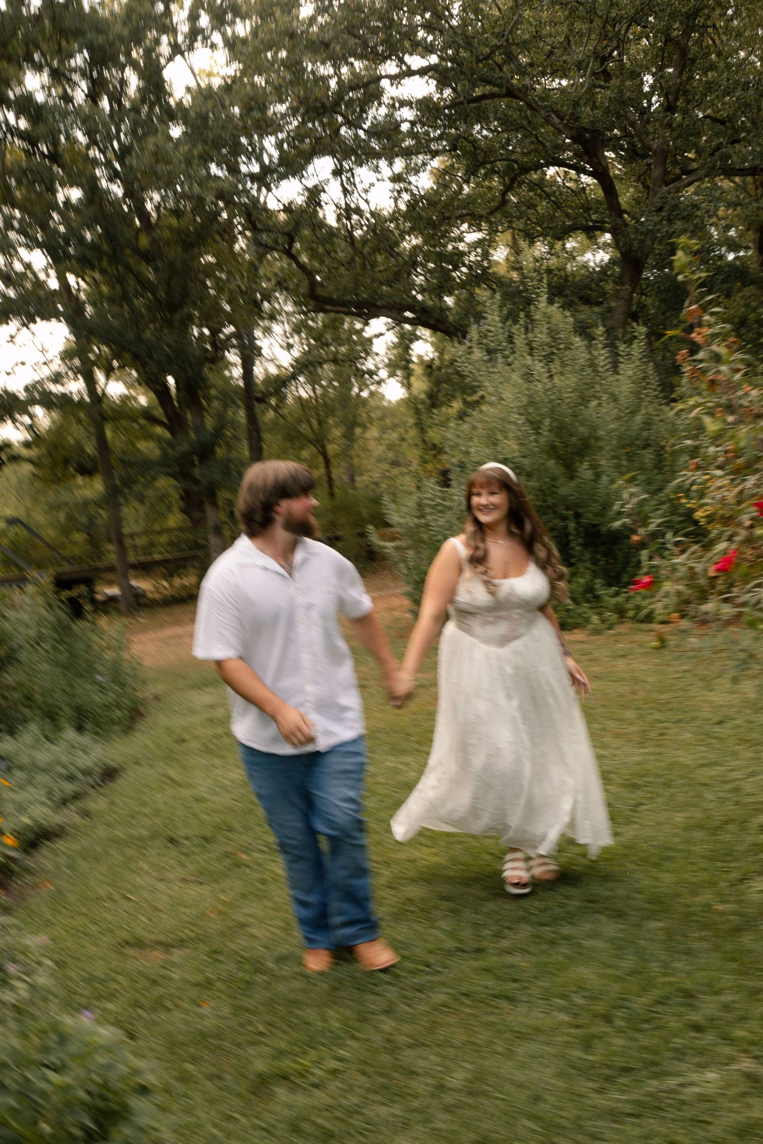 A couple holds hands and walks through a lush green garden, woman wearing a flowing white maternity dress.