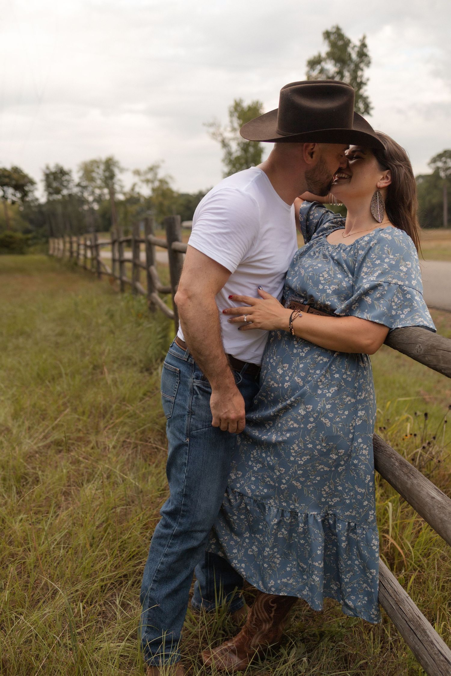 Couple kissing by rustic wooden fence, man in cowboy hat and jeans, woman in blue floral dress, in a pastoral setting.