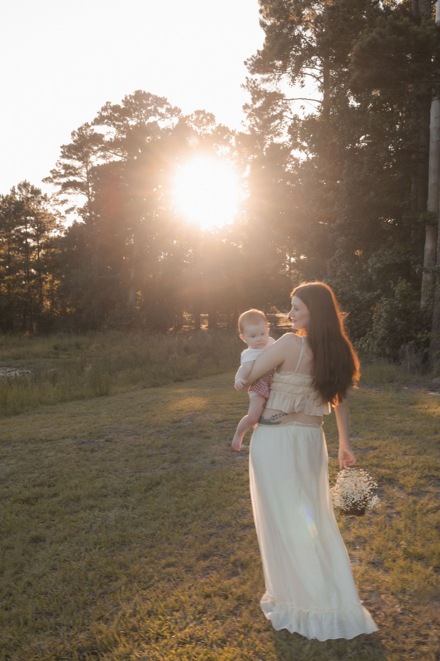 Mother in a white dress holding her baby while standing in a sunlit field at golden hour sunset.