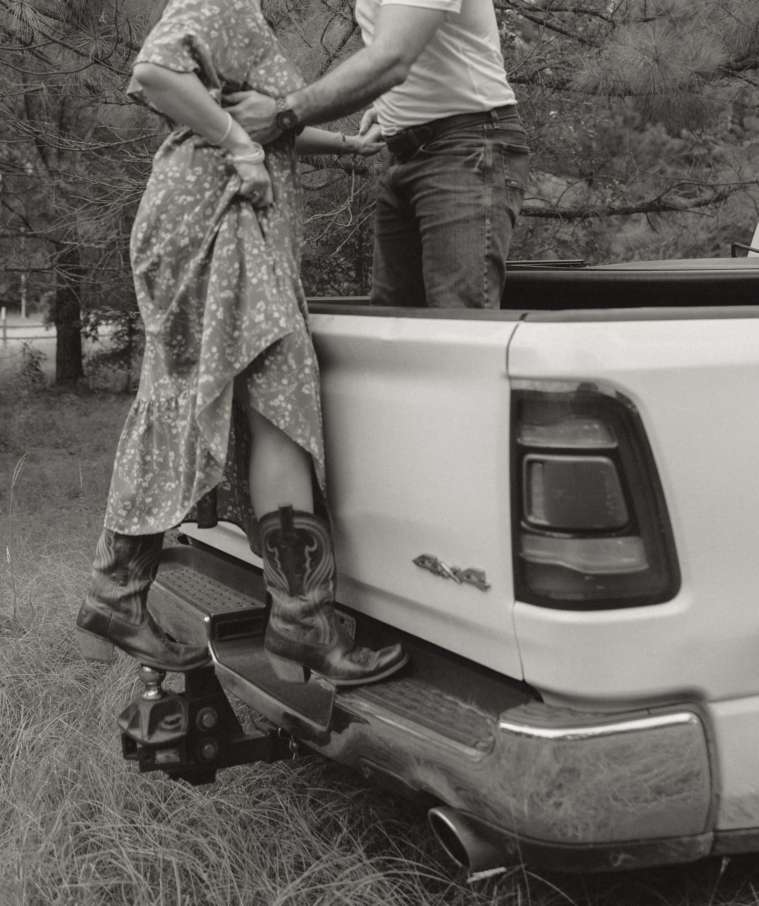 Black and white photo of a couple standing at the back of a pickup truck, woman in floral dress and cowboy boots.