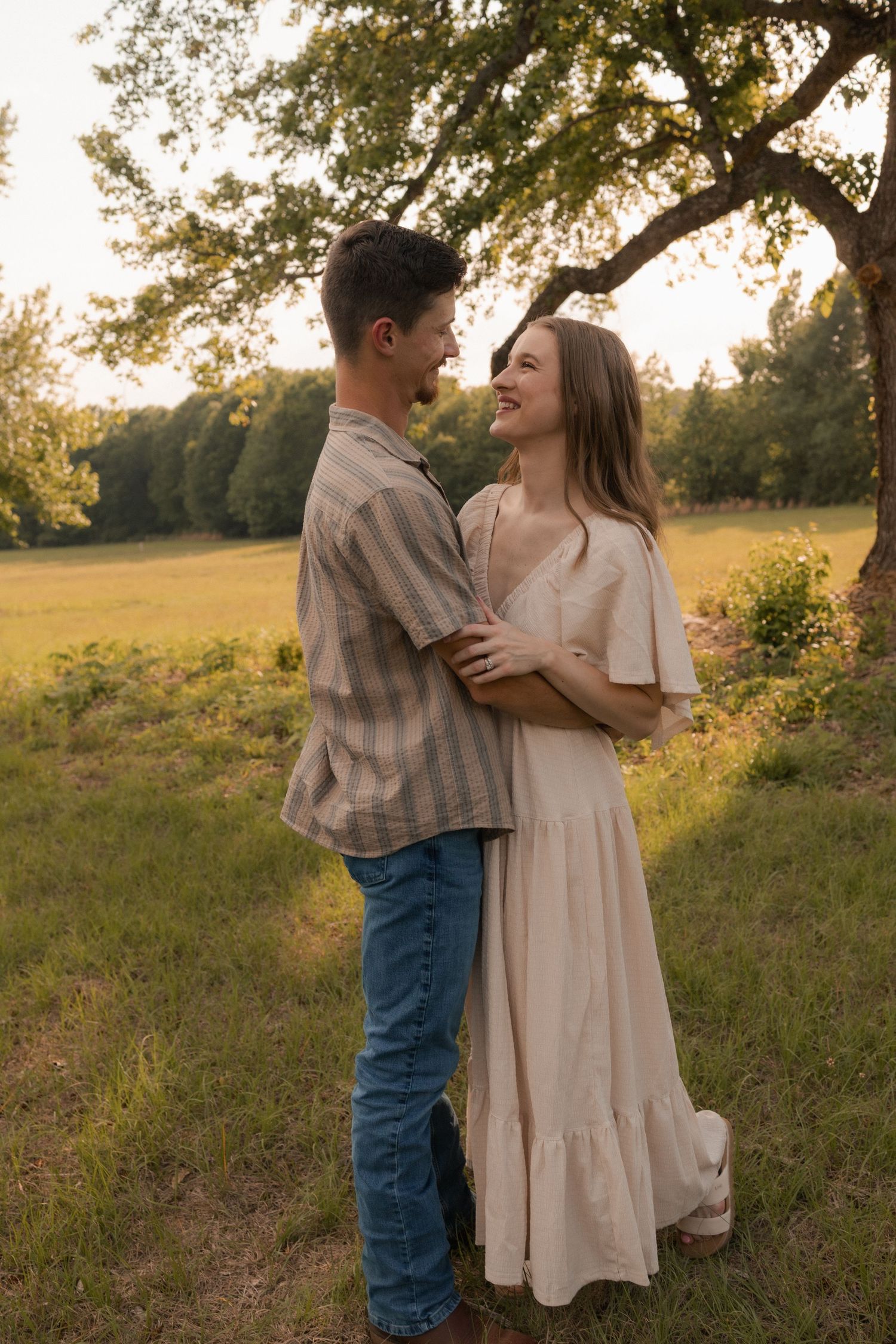 A young couple embraces and smiles at each other in a sunlit green field during golden hour.