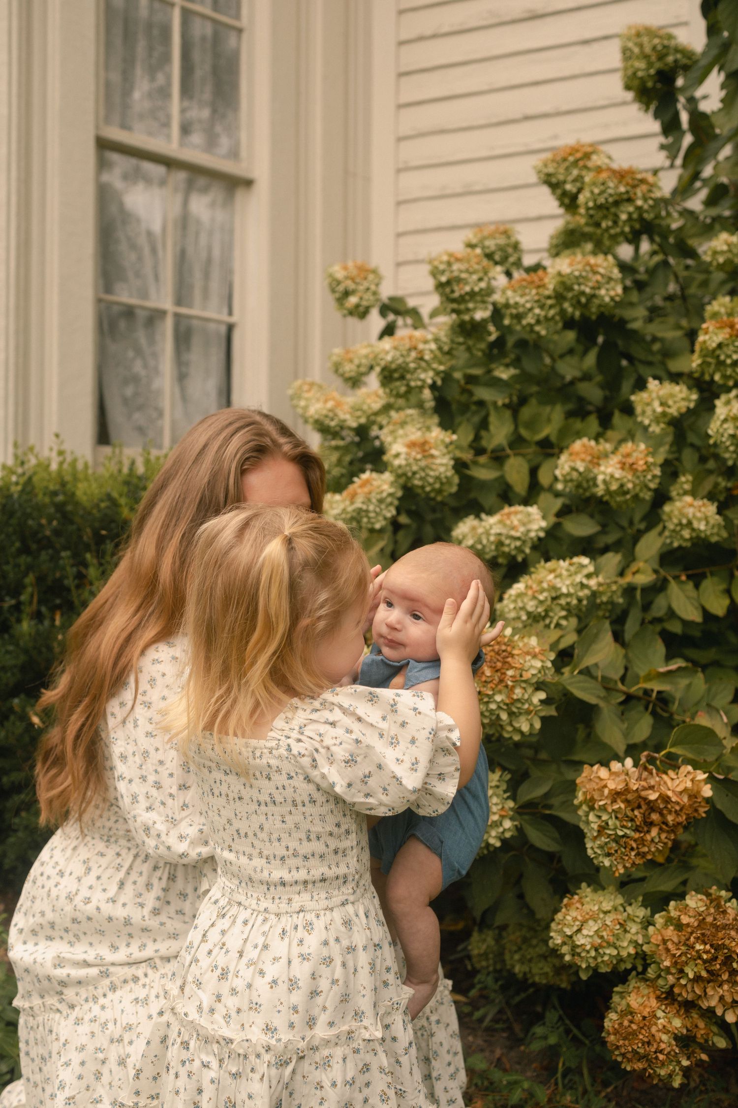 Mother and toddler daughter admiring newborn baby near blooming hydrangea bush outside home.