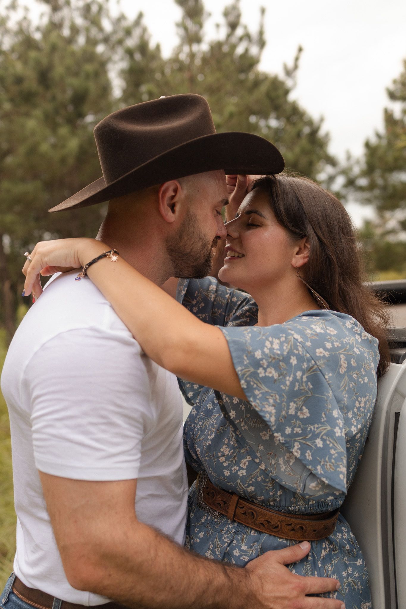 A couple shares a kiss near a truck, man in cowboy hat embracing woman in blue floral dress outdoors.