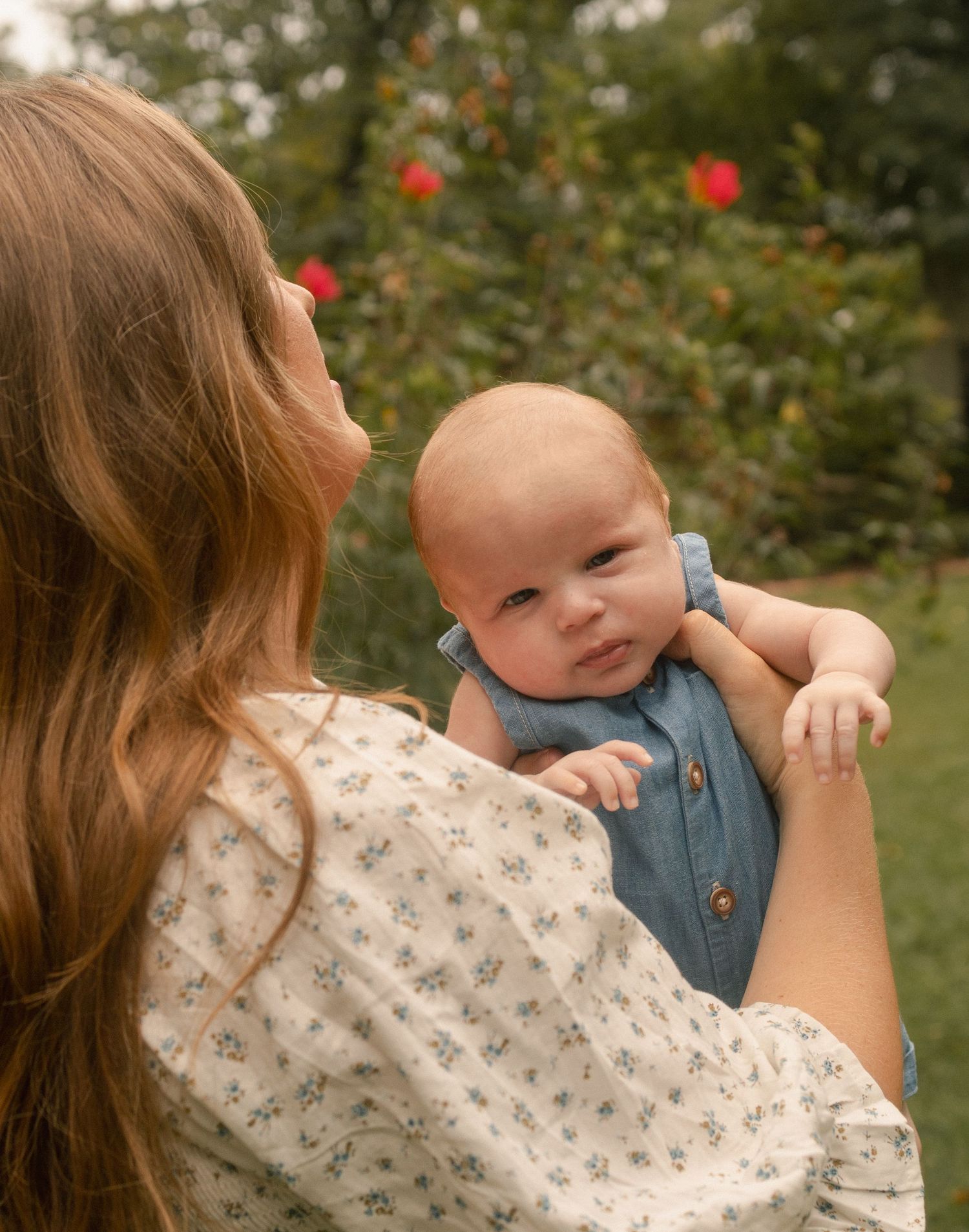 Mother holding newborn baby over shoulder in garden with red roses in background.