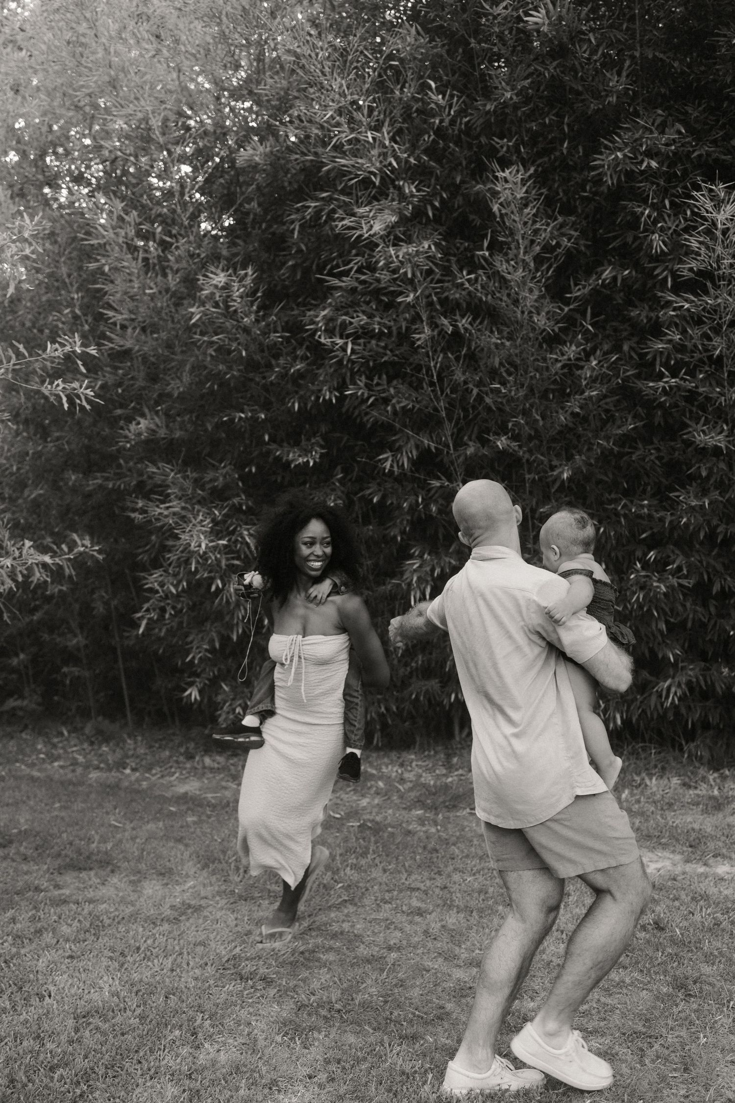 A joyful black and white photo of a smiling couple dancing outdoors, with the man holding their baby.