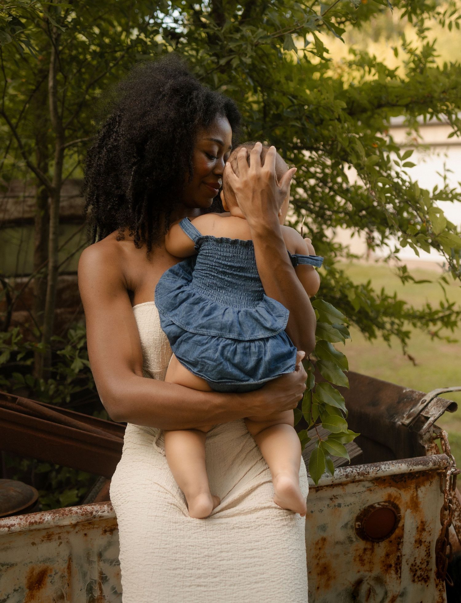 A Black mother in a white dress gently kisses her baby while standing near a rustic vintage truck.