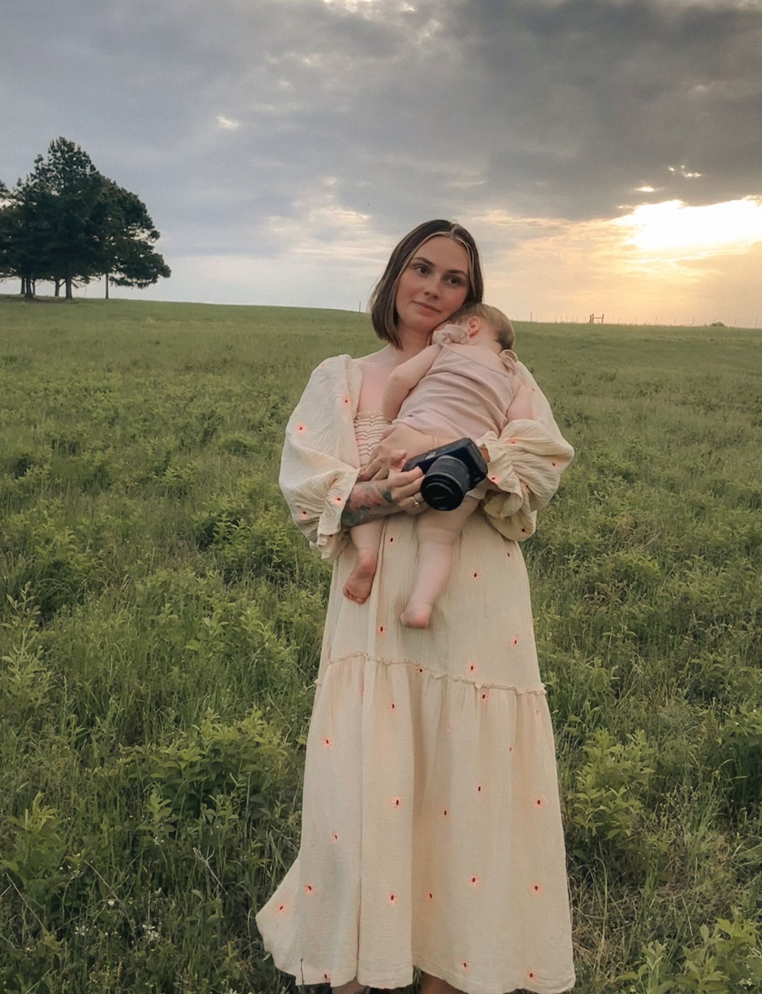 Mother in cream polka dot dress cradling newborn baby in lush green field at golden sunset.