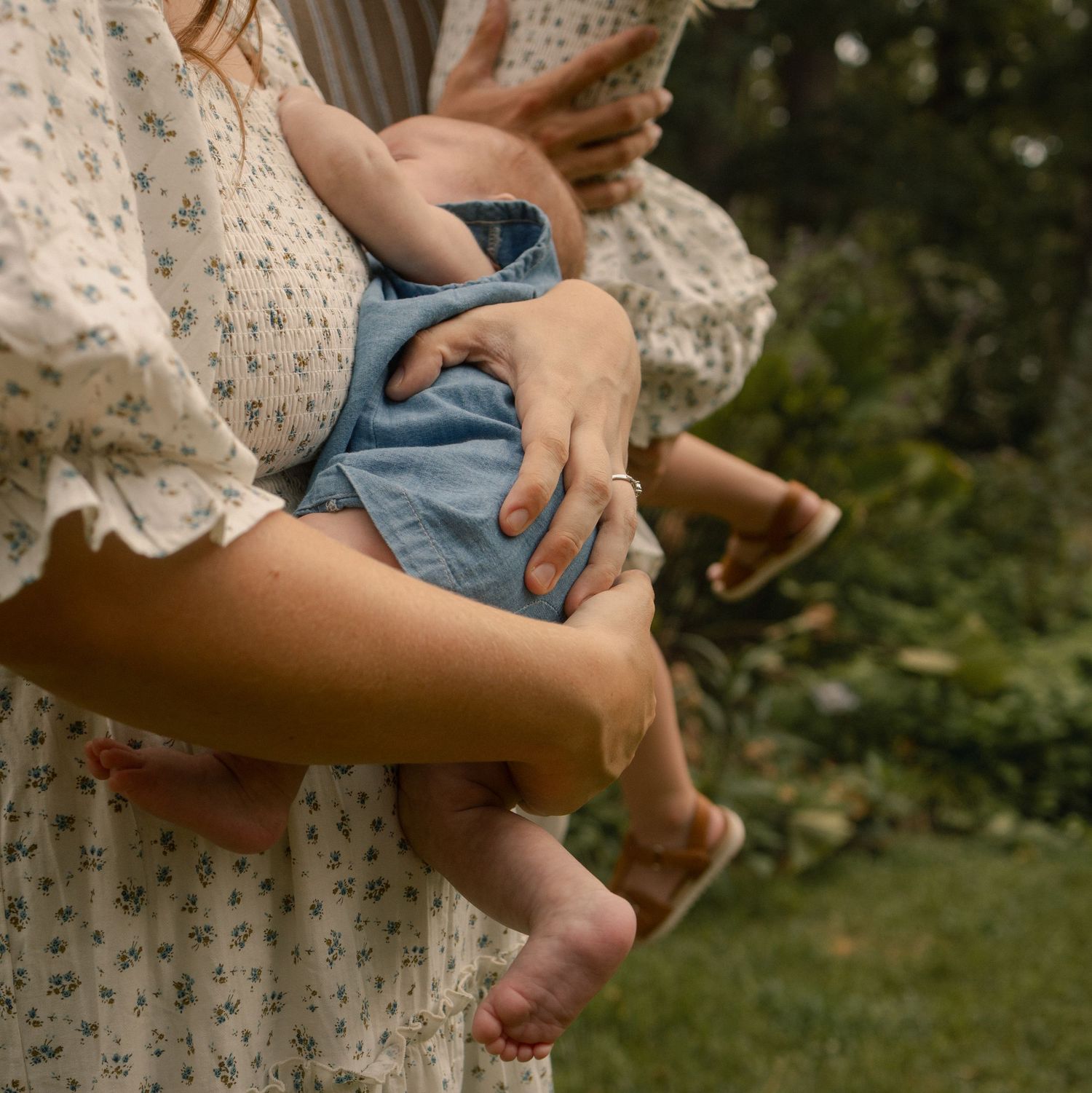 Close-up of mother holding baby in blue romper with bare feet in garden setting.