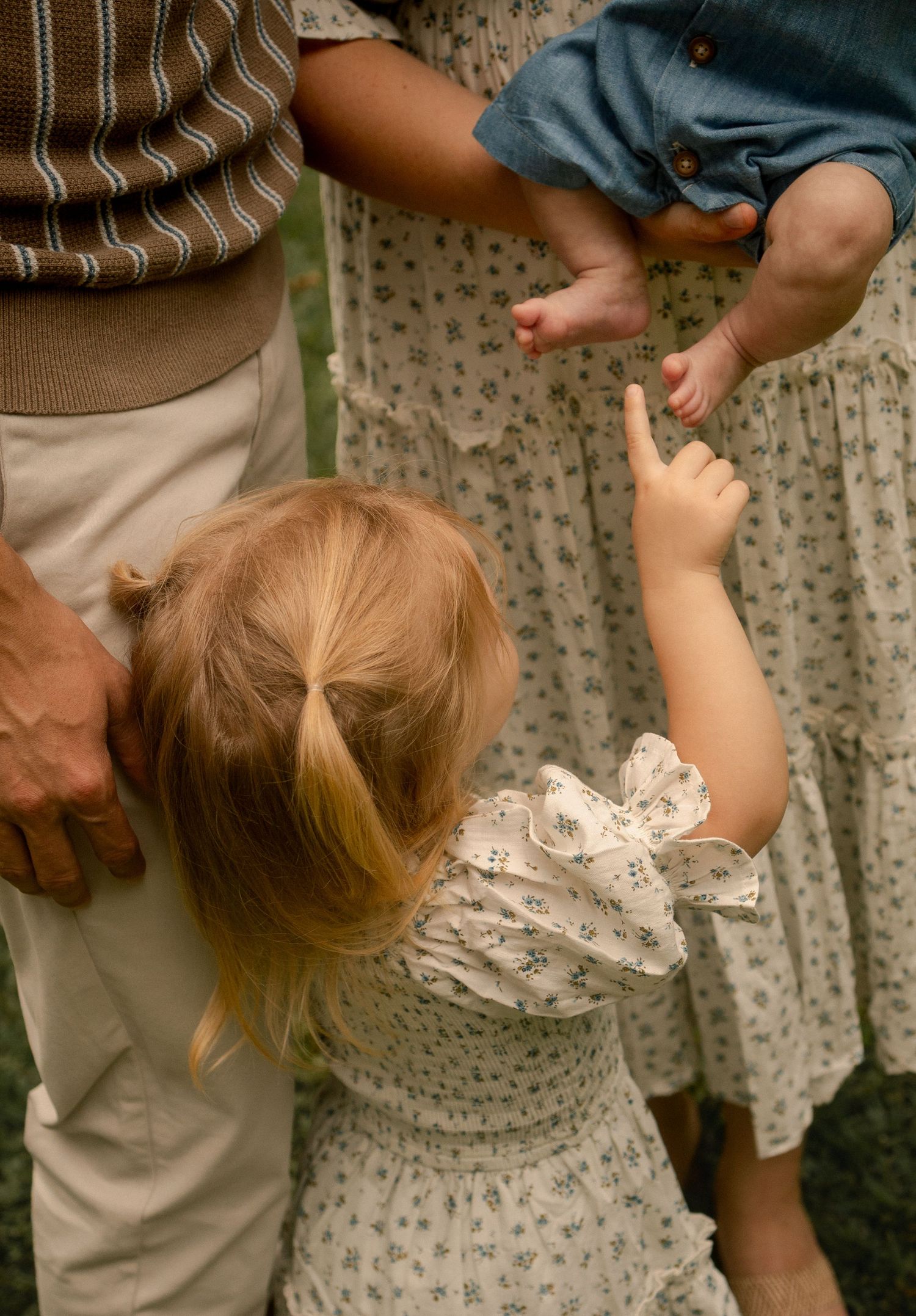 Young toddler girl in floral dress pointing at baby sibling held by parents outdoors.