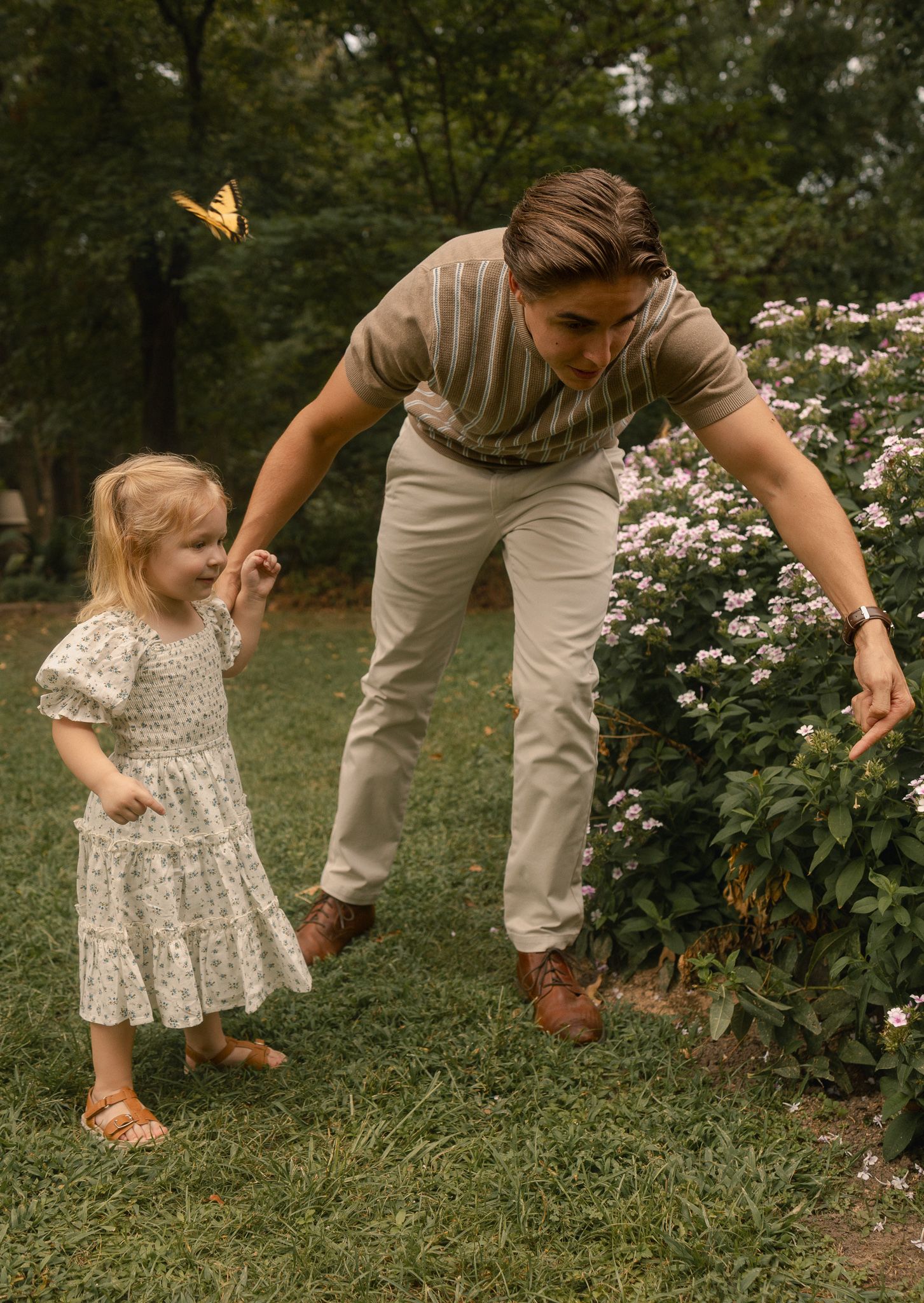 A father and young daughter discover a butterfly near blooming flowers in a lush garden setting.