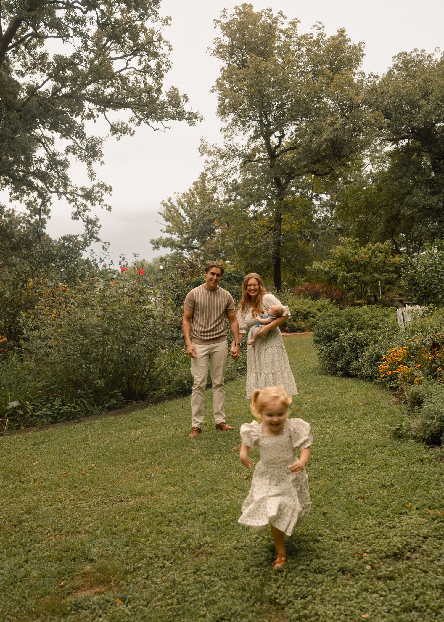 A toddler runs toward the camera while her parents walk behind her on a lush green garden path.