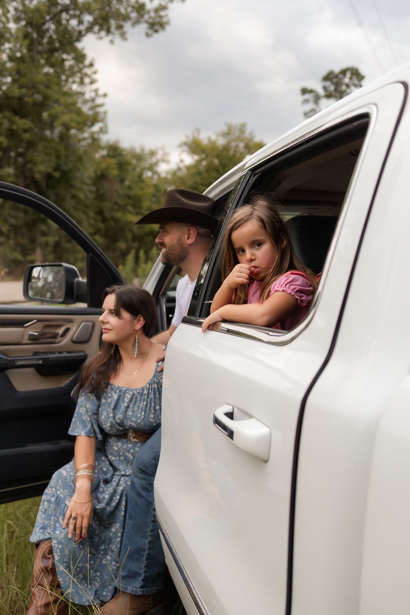 Family of three poses by a white pickup truck, with a girl leaning out the window and parents standing outside.