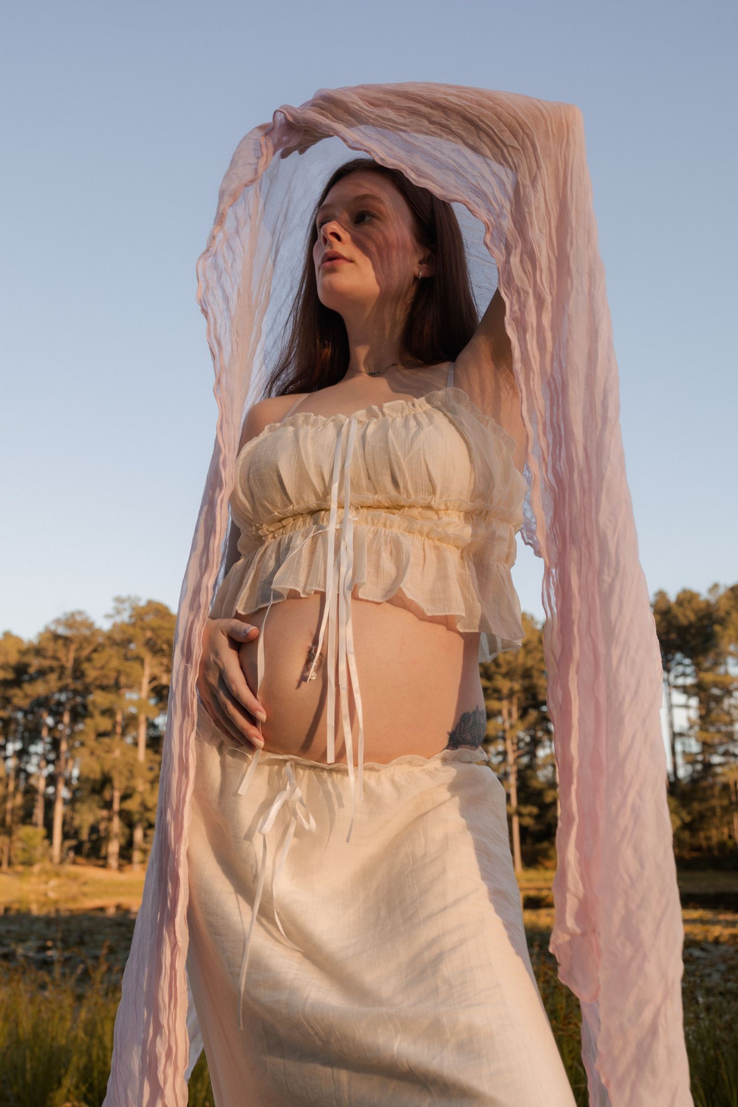 Pregnant woman in a white crop top and skirt holding her belly with pink fabric flowing above outdoors.
