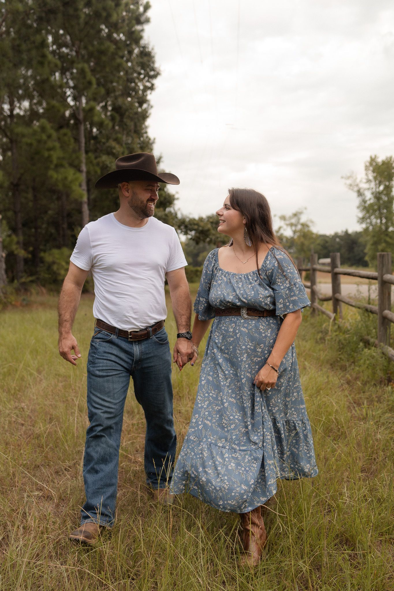A couple in country attire hold hands walking through a grassy field, man in cowboy hat and woman in blue floral dress.