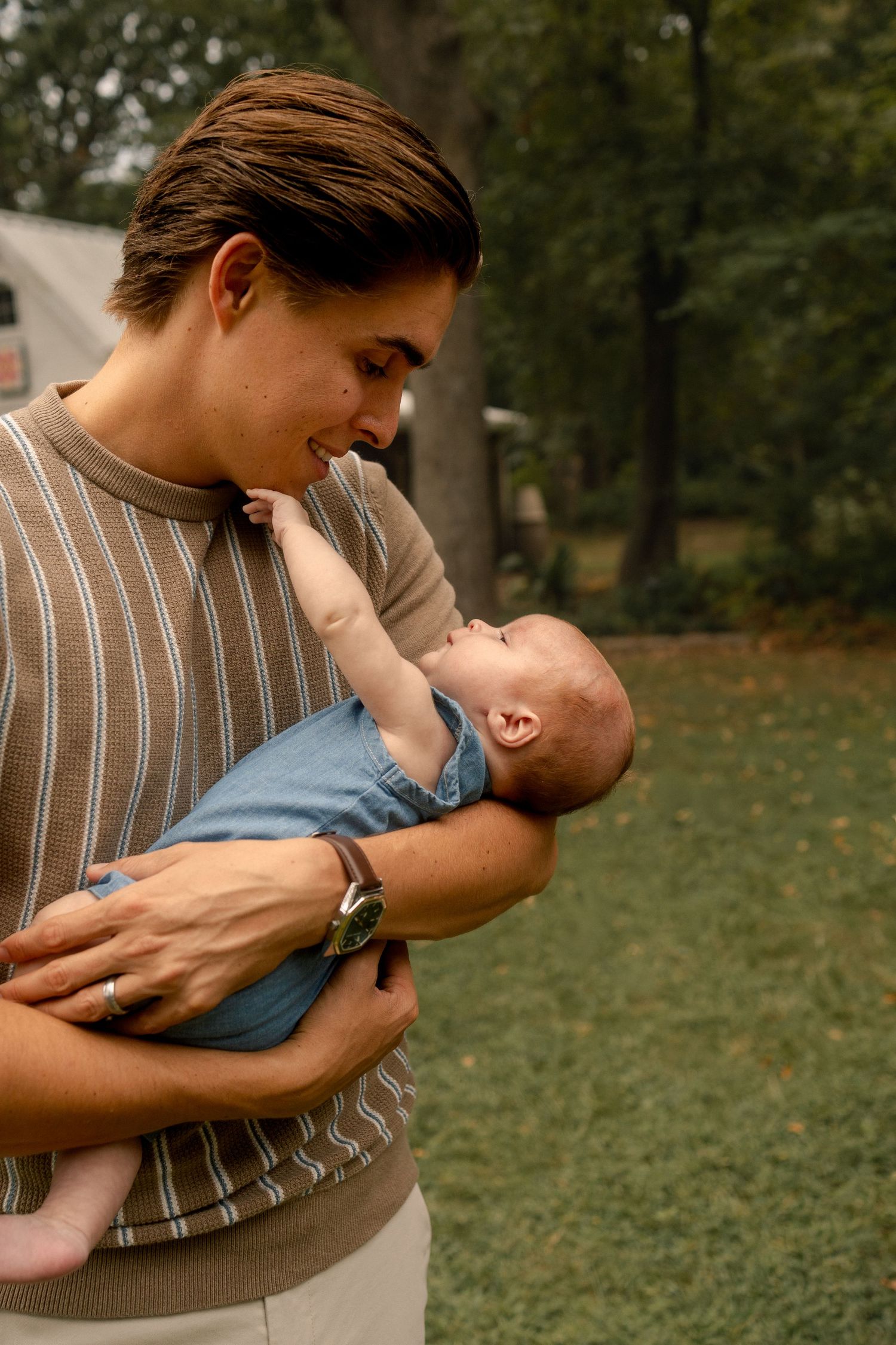 Smiling parent in striped sweater holding and gazing at happy baby in outdoor setting.