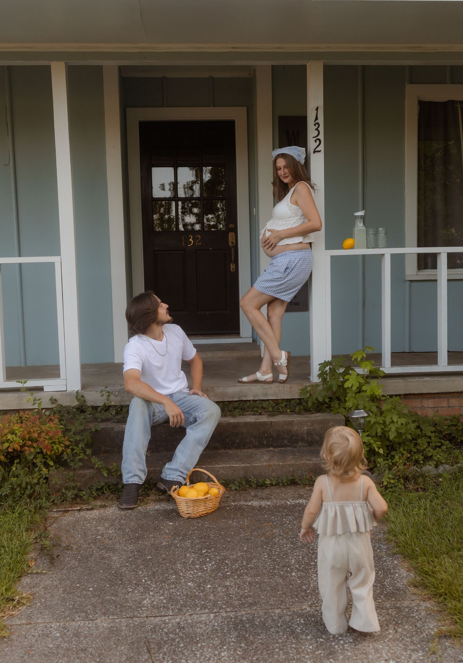 Pregnant woman on porch steps with partner and toddler outside a light blue house.
