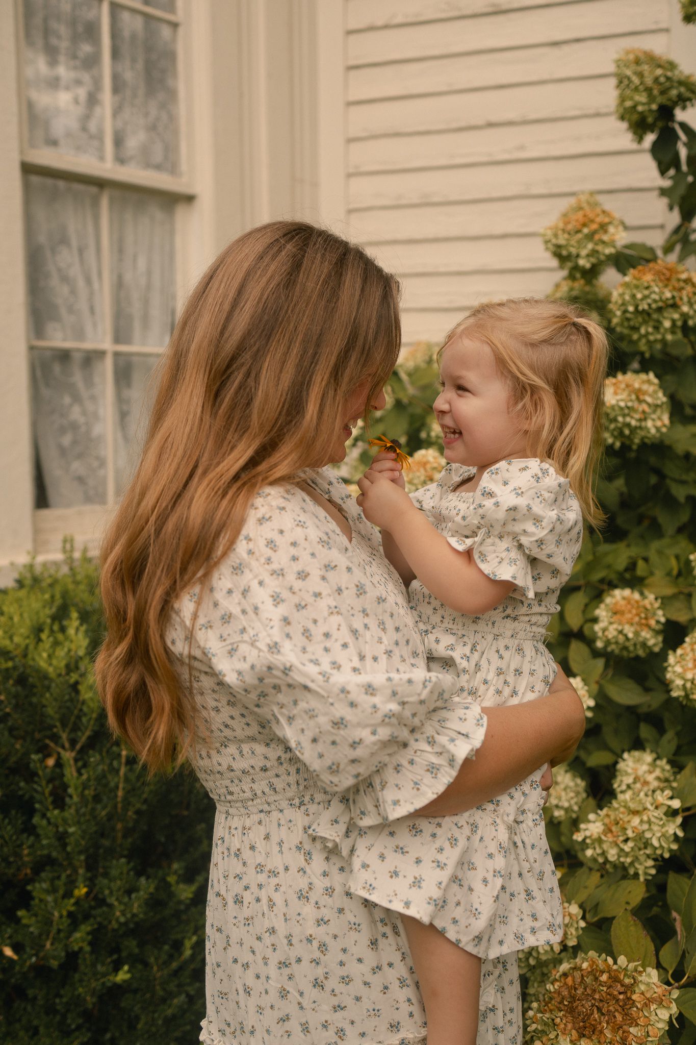 A mother with long auburn hair holds her smiling toddler daughter near hydrangea bushes by a white house.