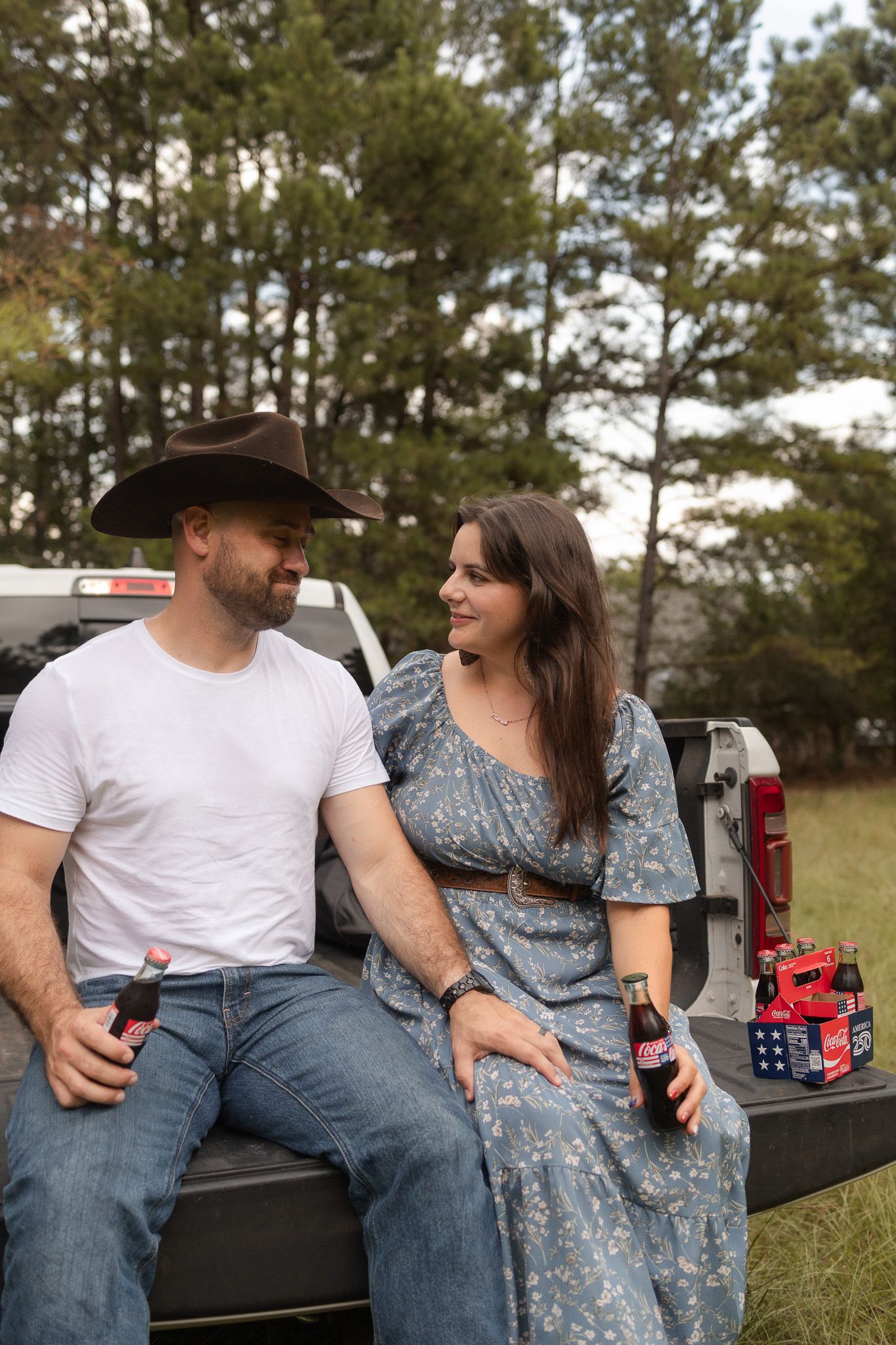 Couple sitting on a truck tailgate holding Coca-Cola bottles, man wearing a cowboy hat outdoors.