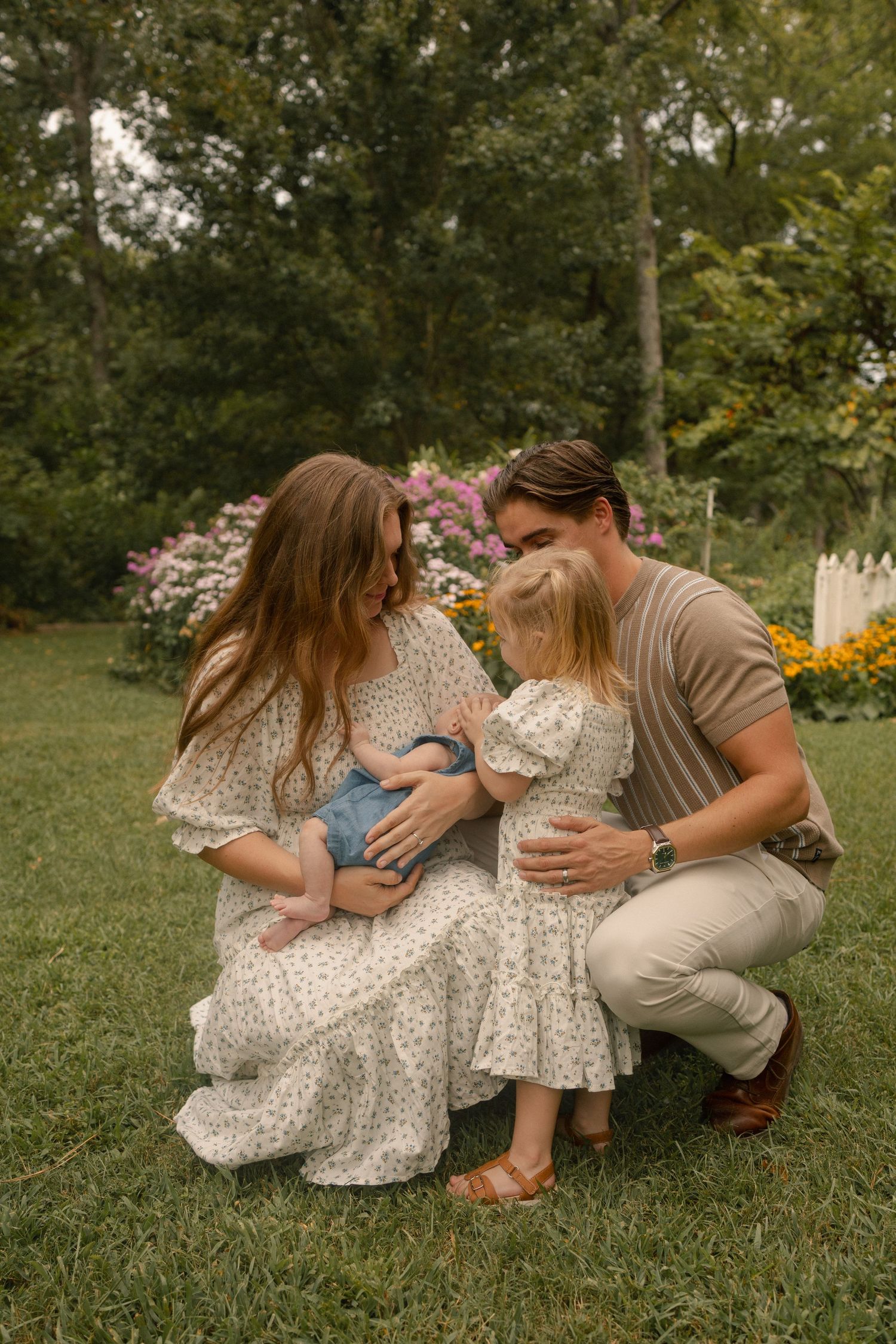 A family of four gathers closely on a garden lawn, parents and toddler admiring their newborn baby.