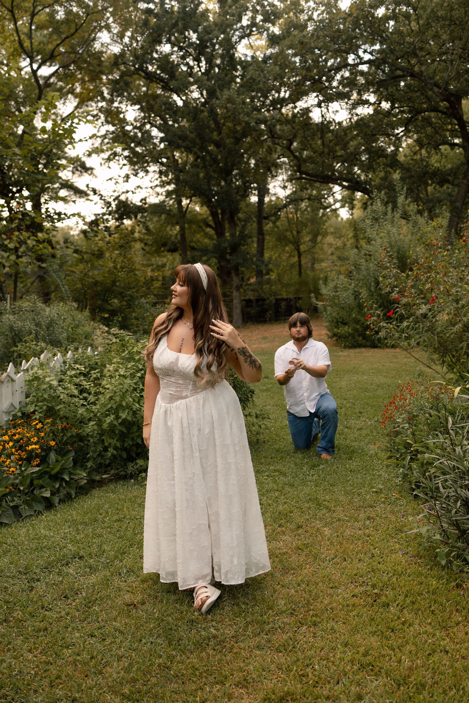 Woman in a white dress walking in a garden while a man kneels behind her in a proposal moment. Located in Cameron NC.