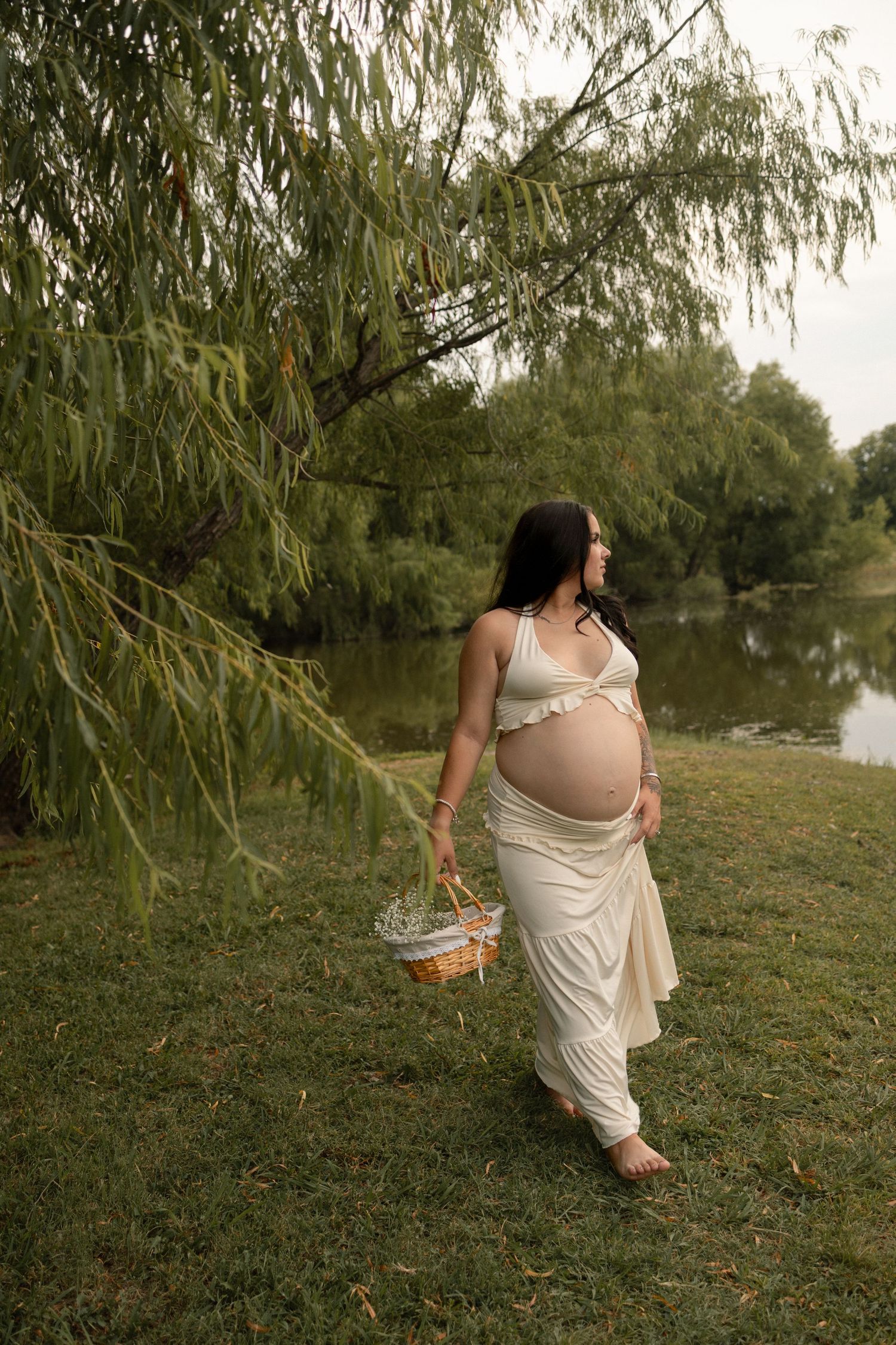 Pregnant woman in white outfit carrying wicker basket under weeping willow by lake.