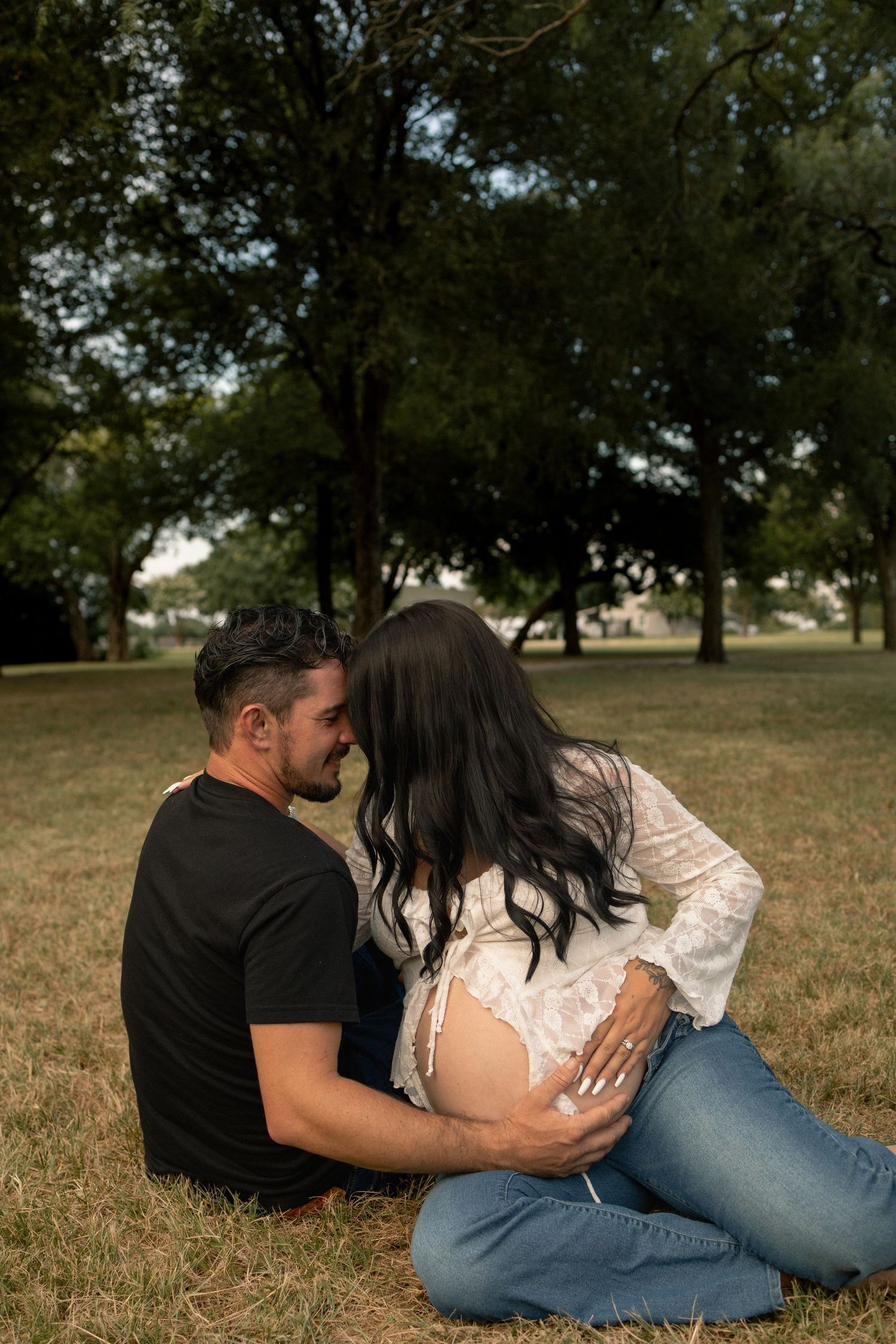 A couple sits intimately on grass in a park, man tenderly holding pregnant woman's baby bump.