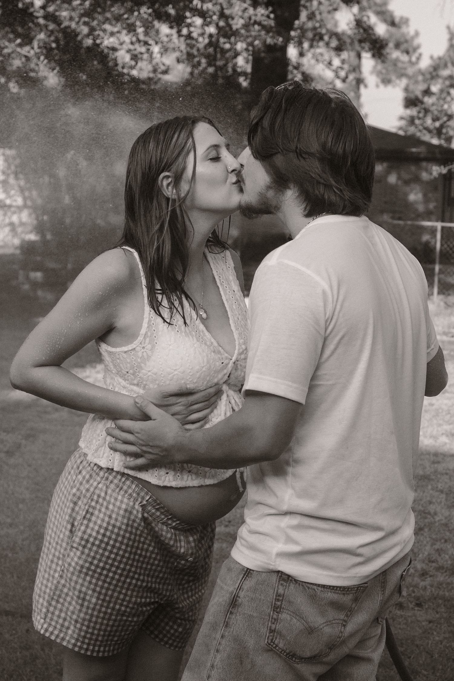 Black and white photo of couple kissing in the rain, man cradling pregnant womans belly.