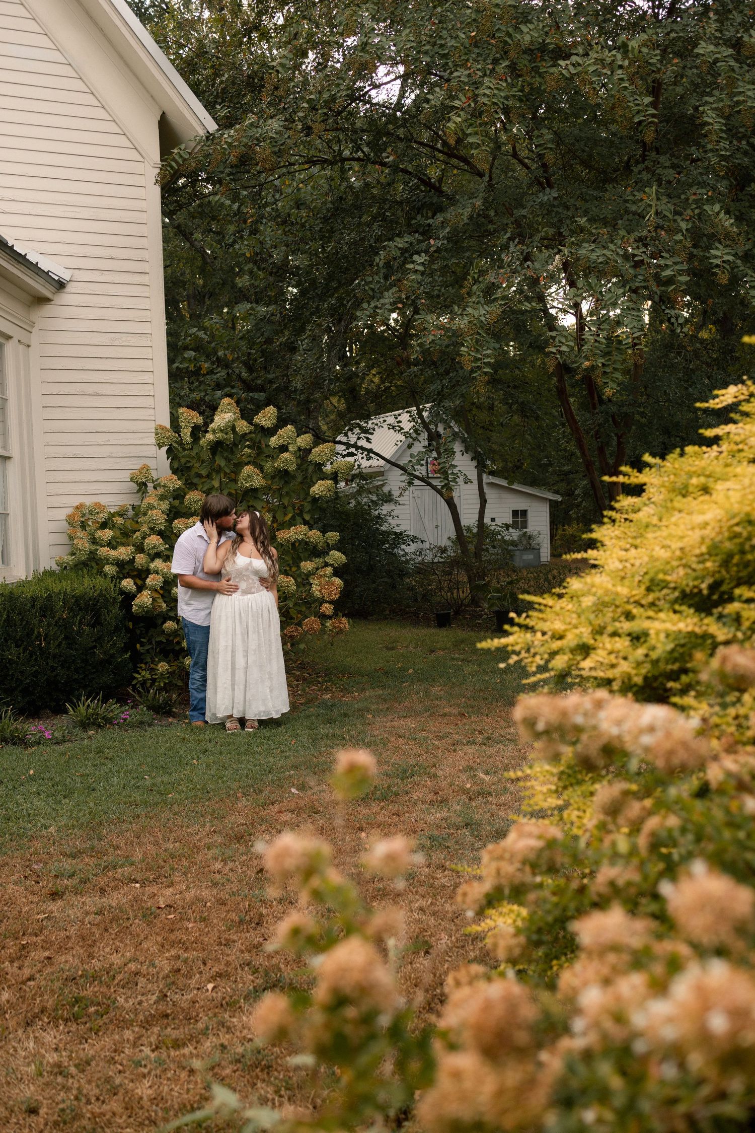 Couple sharing a romantic moment in a lush garden beside a white house, surrounded by blooming hydrangeas and yellow shrubs.