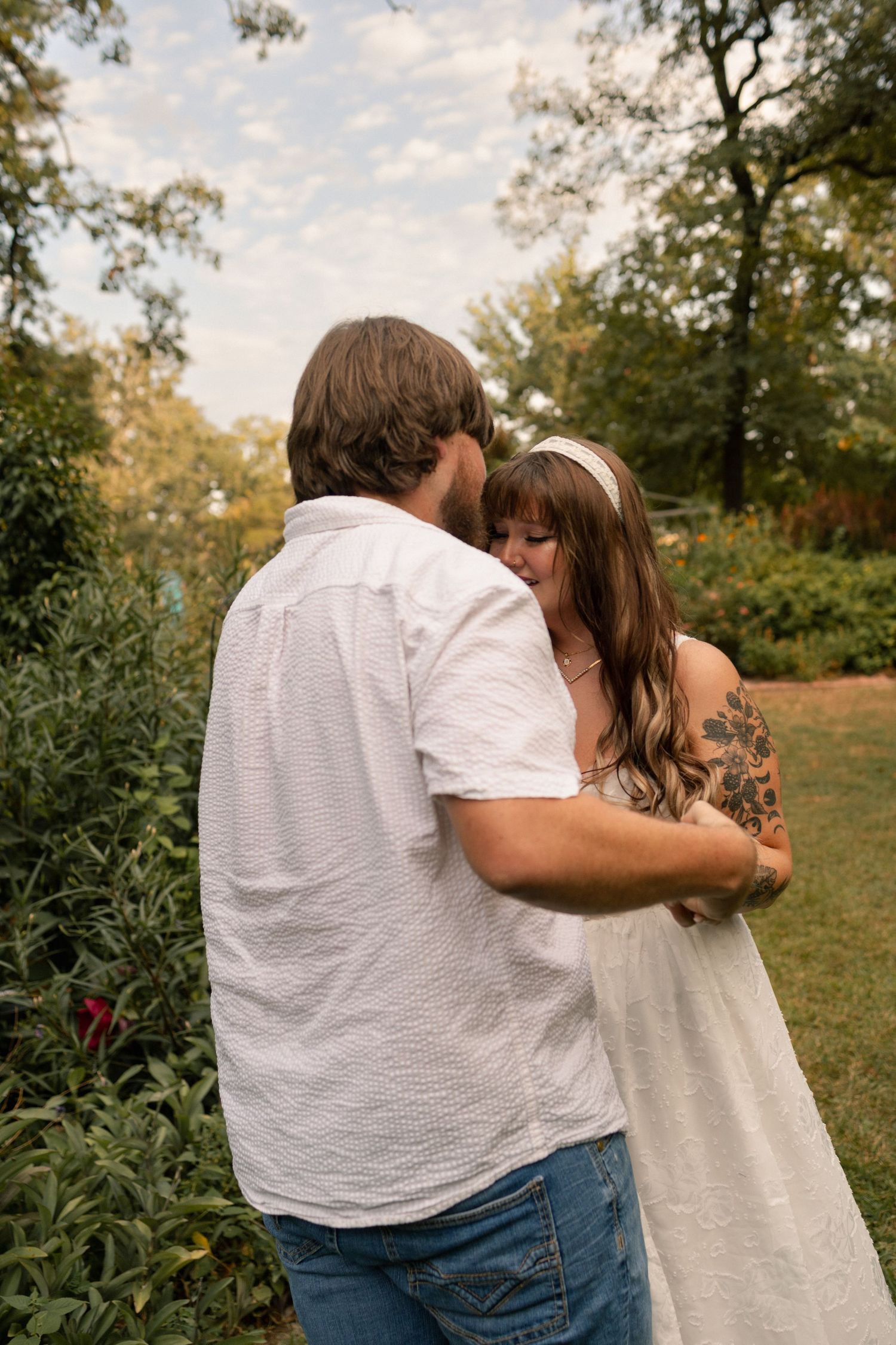 A couple shares a tender embrace in a lush garden, woman in white dress with a floral tattoo.