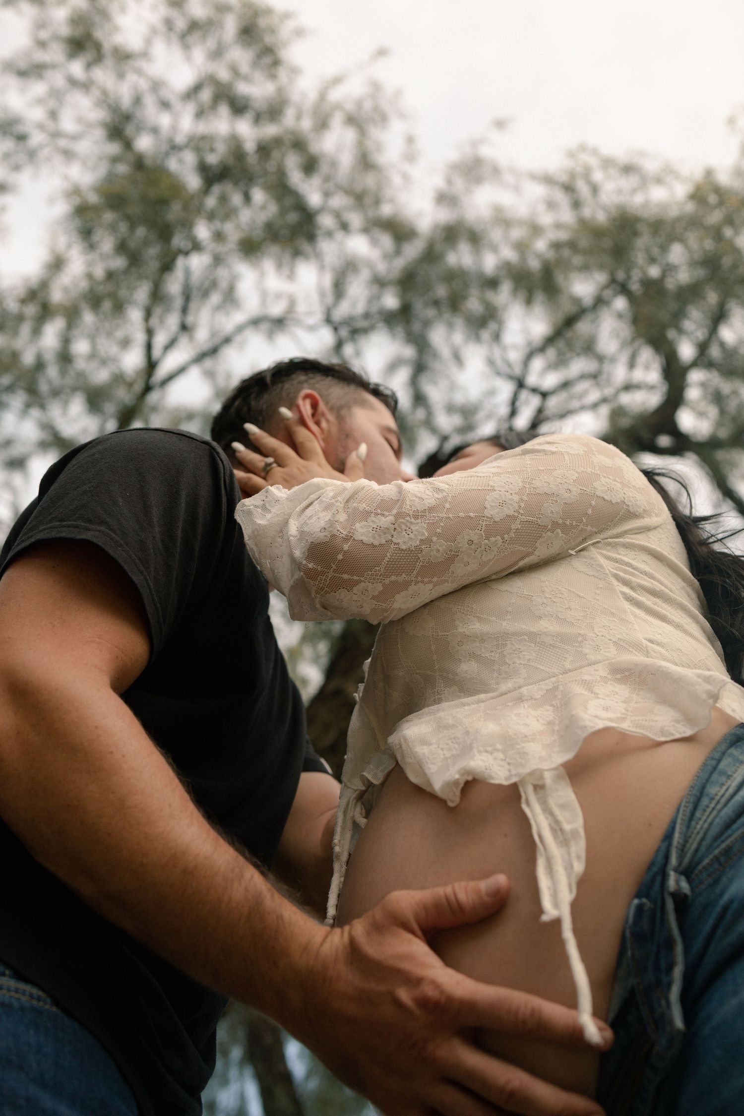 A man tenderly holds a pregnant woman's belly while they share an intimate kiss outdoors.