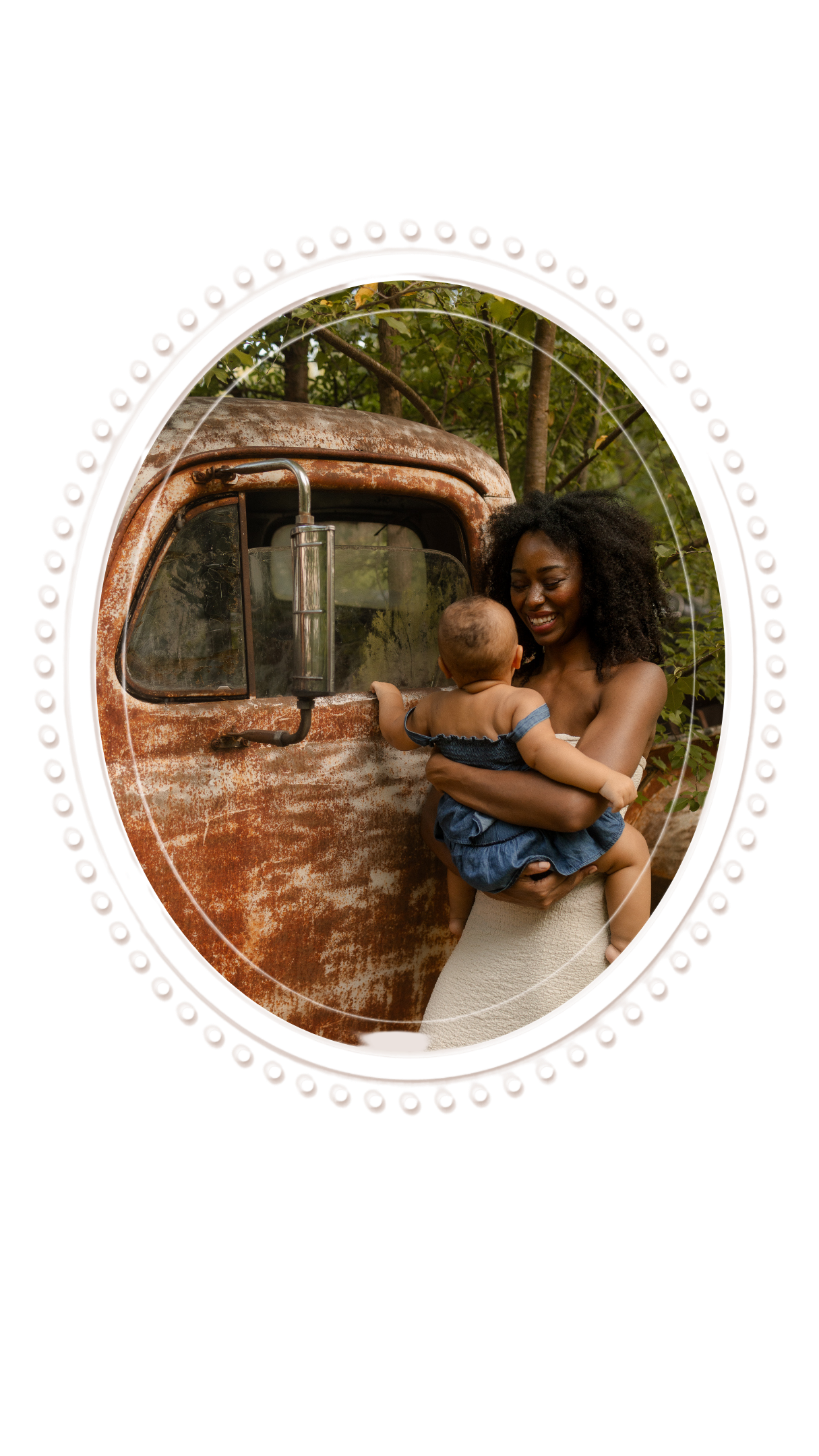 Joyful mother cradling her infant beside a weathered rusty truck surrounded by lush green trees.