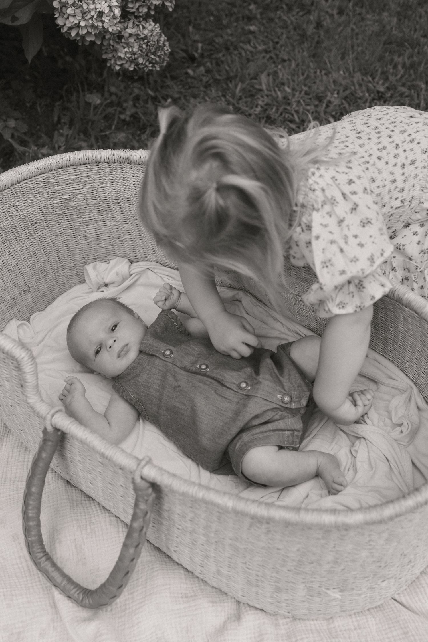 Black and white photo of a toddler girl leaning over a wicker bassinet to look at a newborn baby outside.