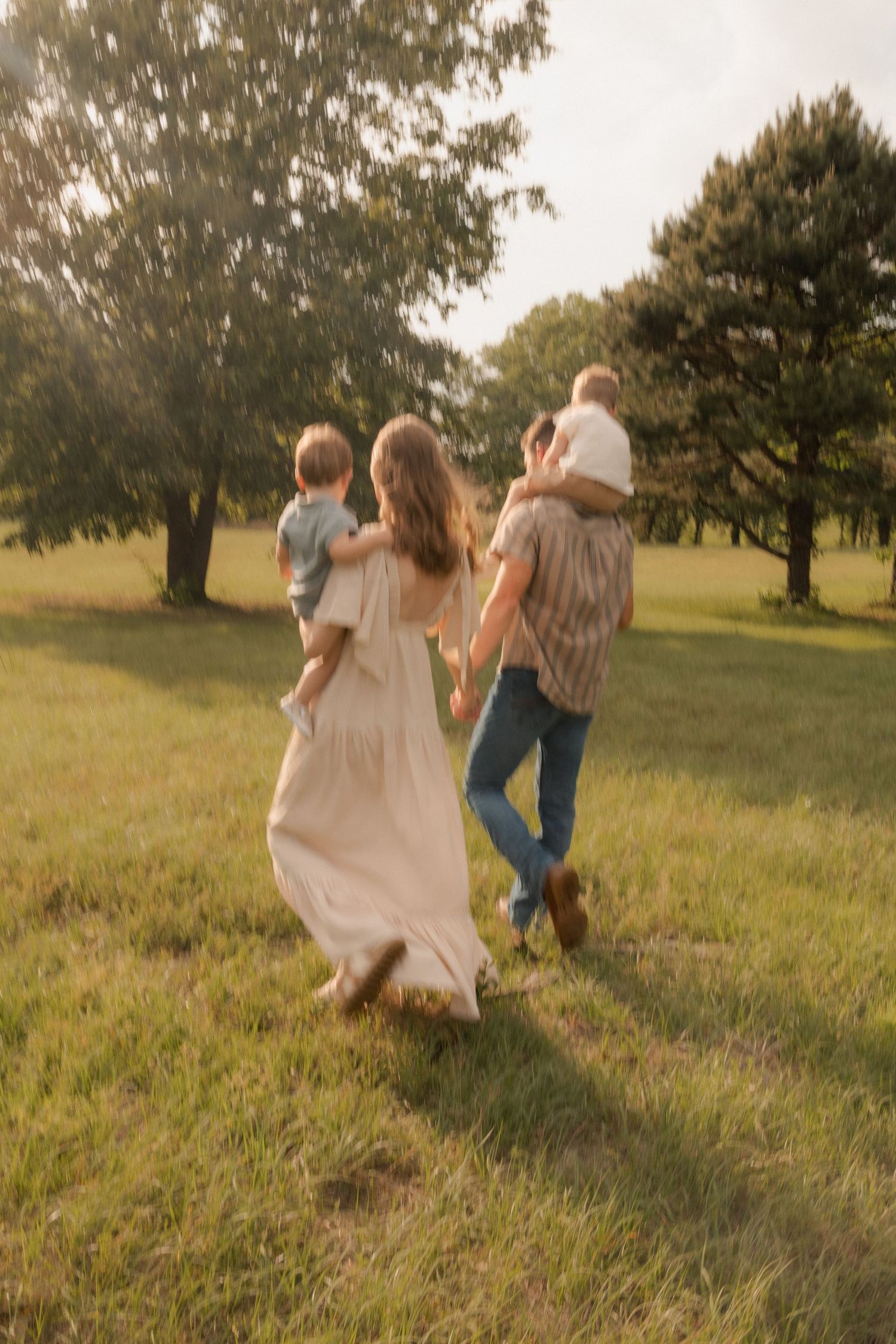 Family walking through sunny green field, mother in white dress carrying toddler, father with child on shoulders.