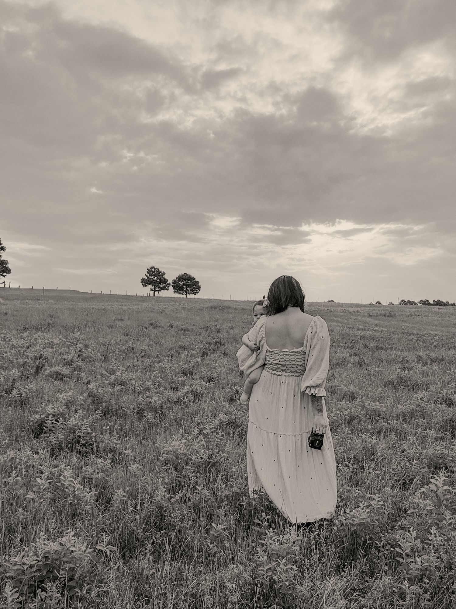 Black and white photo of woman in white dress photographing open field with dramatic cloudy sky and distant trees.