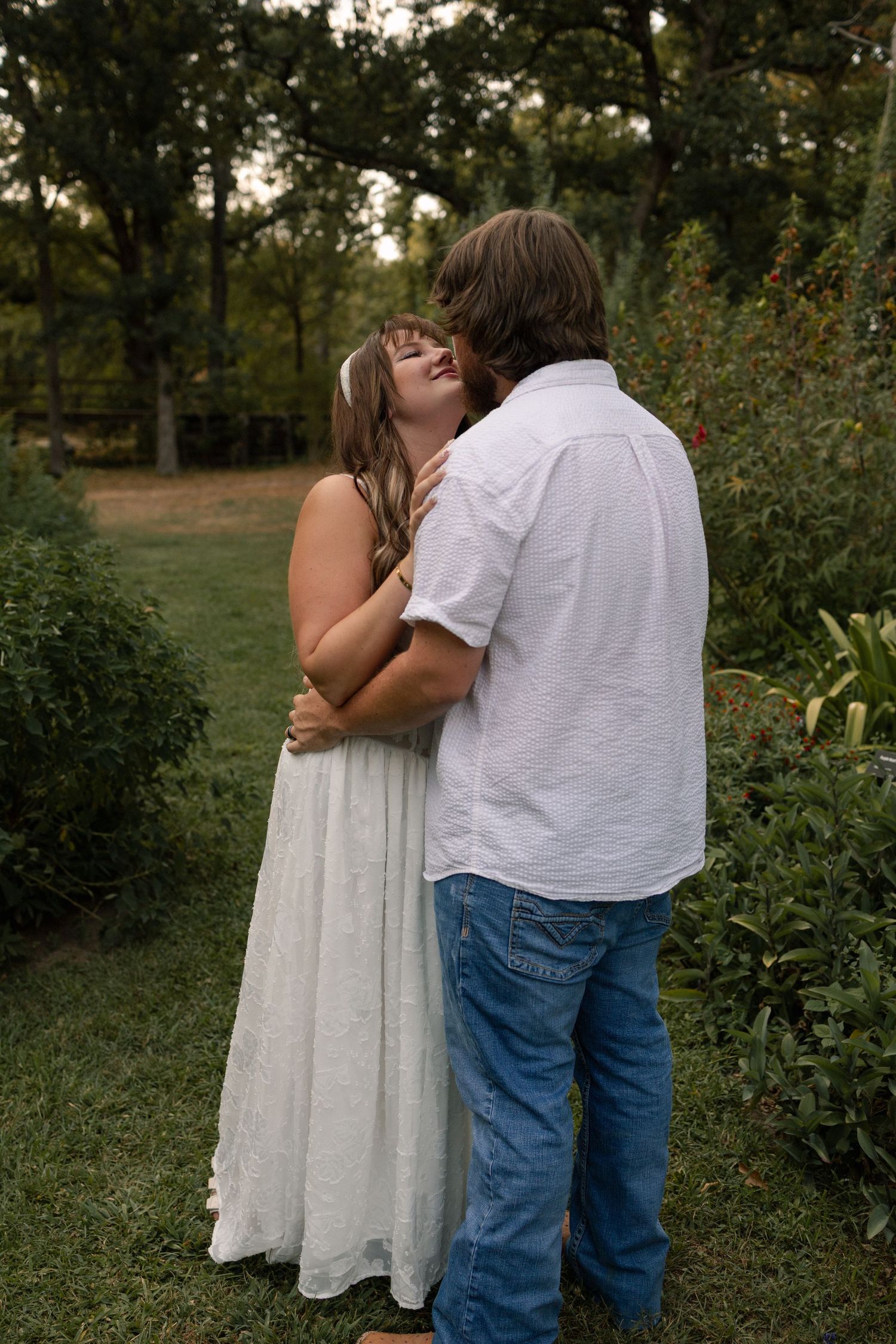 Couple embracing and kissing in a lush green garden, woman in white dress and man in white shirt.