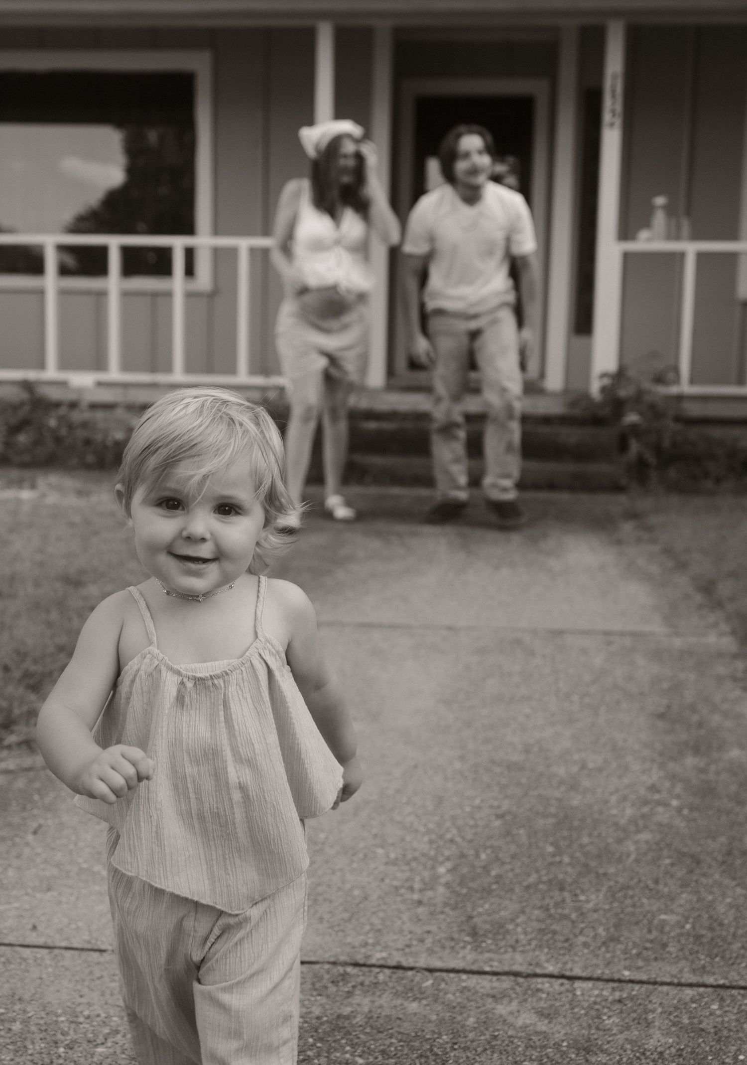 Black and white photo of a toddler girl running toward the camera with her pregnant mother and father standing on a house porch.